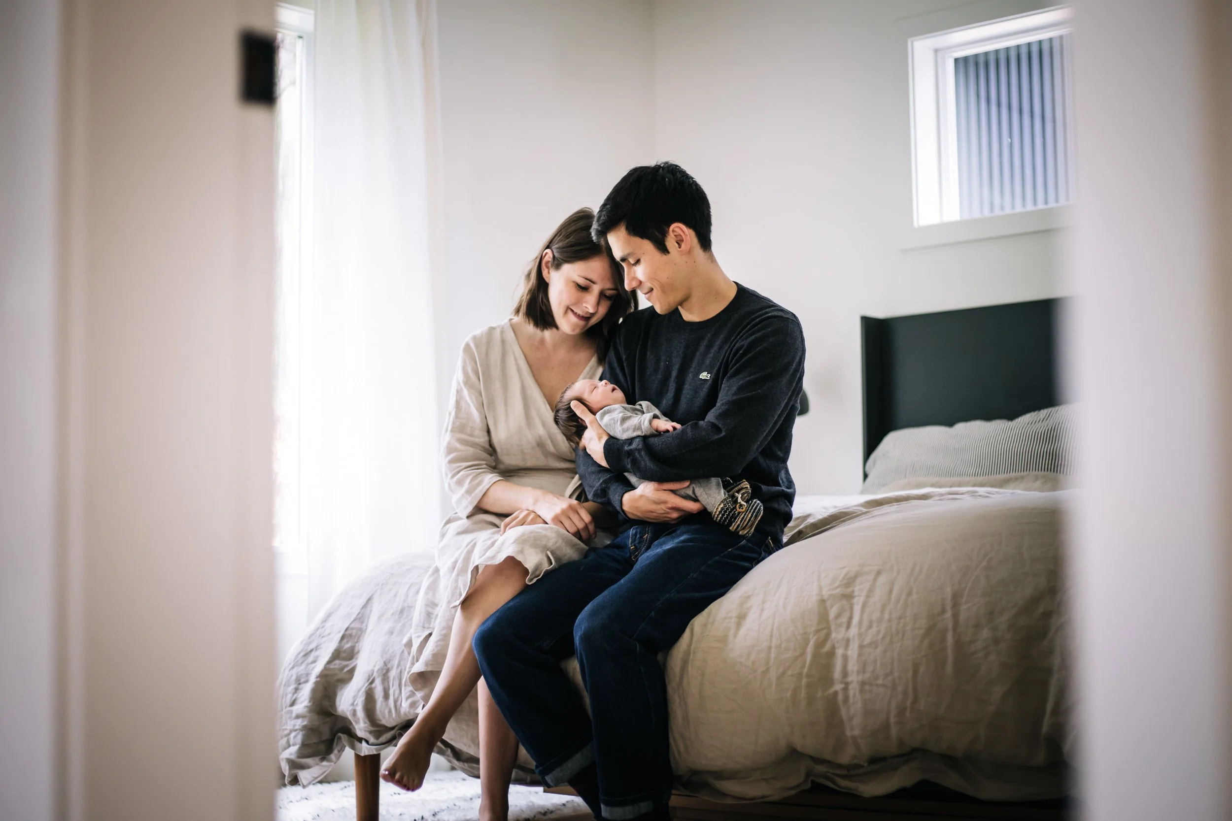 Couple sitting on bed holding a baby in a bright, cozy room.
