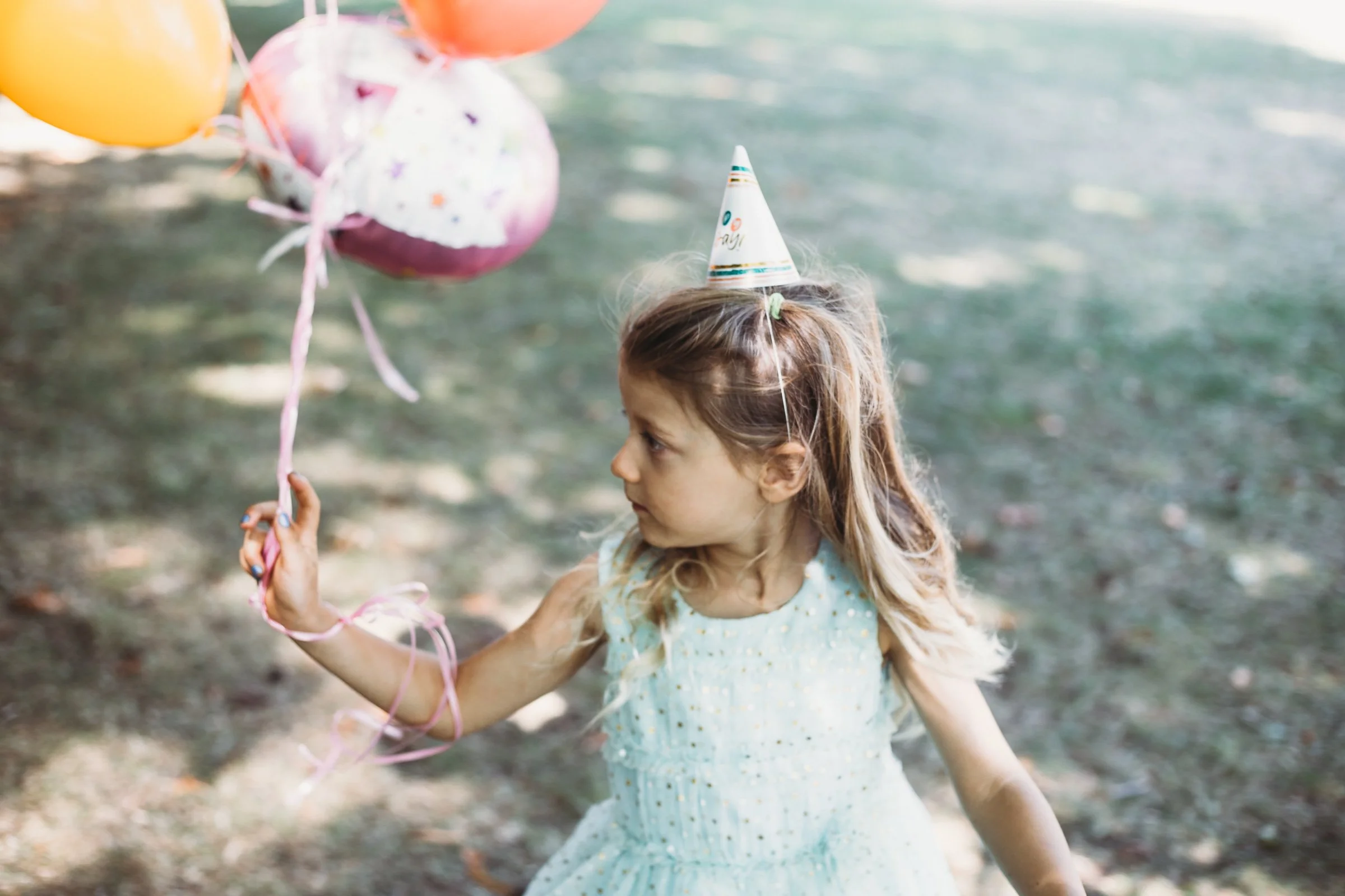 Young girl with balloons and party hat outdoors at a celebration.