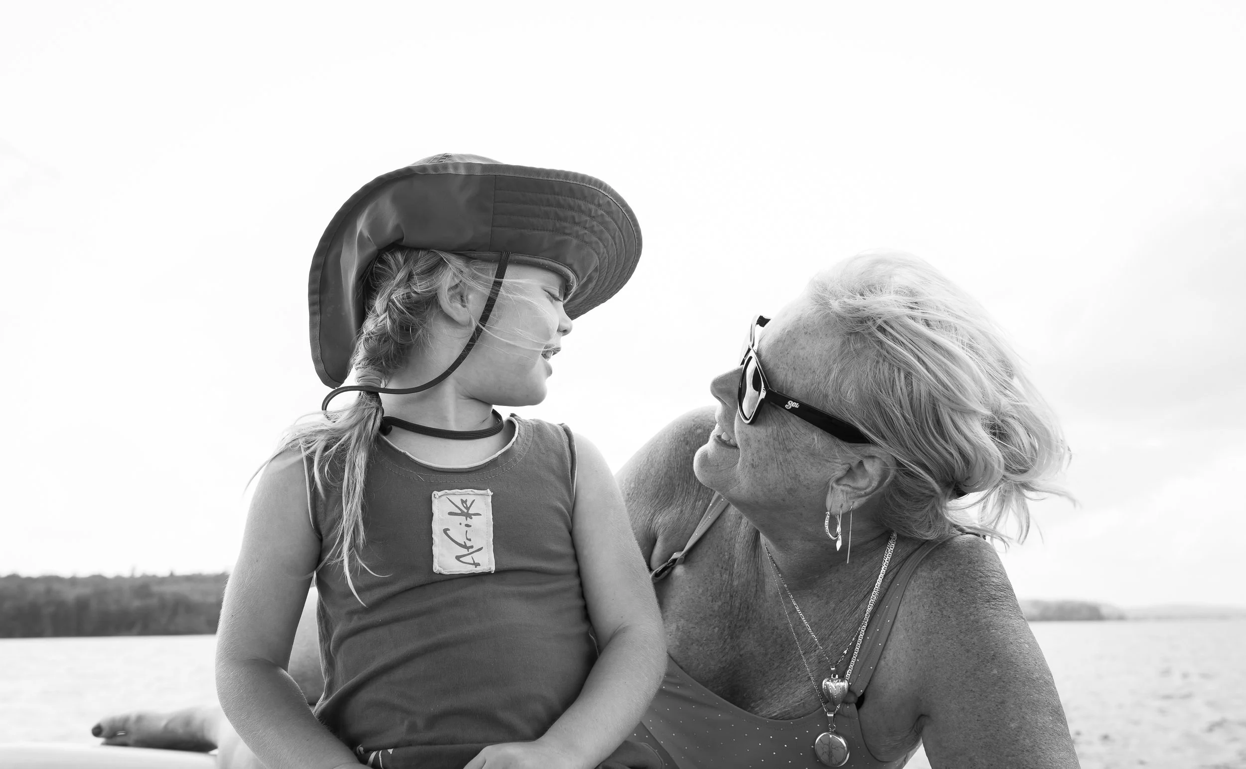 Black and white photo of a child in a sun hat and a woman wearing sunglasses, smiling at each other outdoors near water.