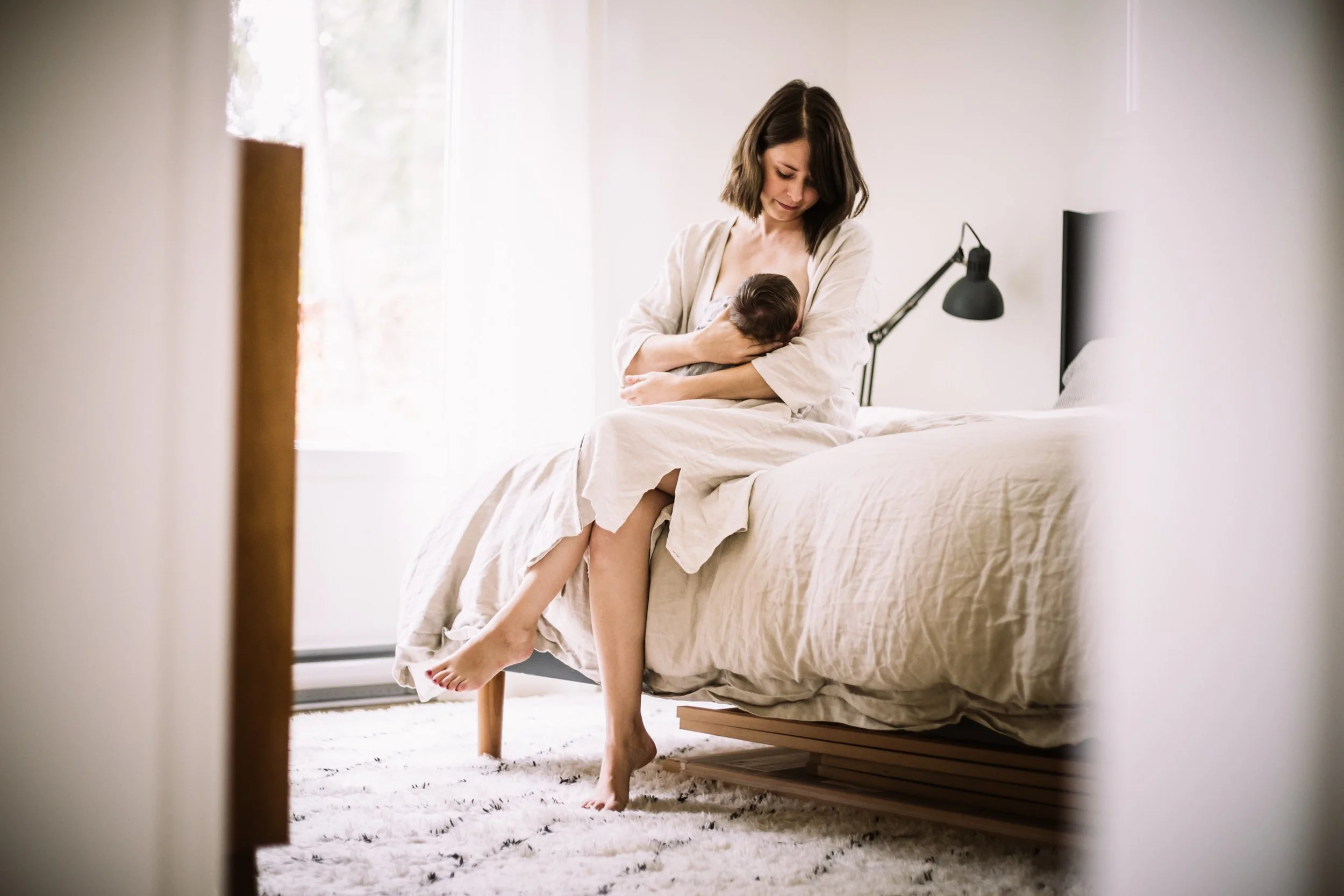 Mother sitting on bed breastfeeding newborn in a bright bedroom.