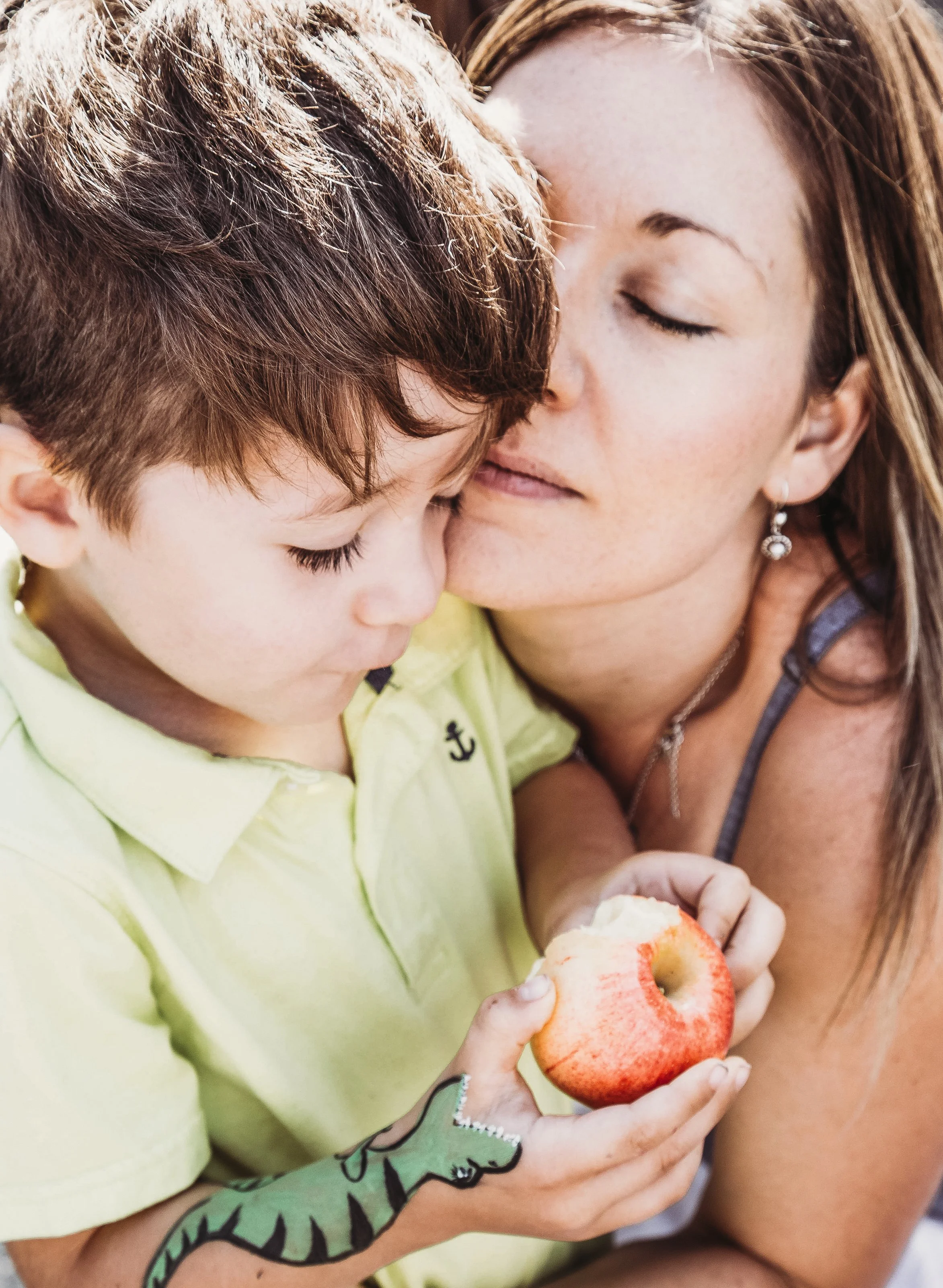 Mother embracing child holding apple with dinosaur face paint on arm.