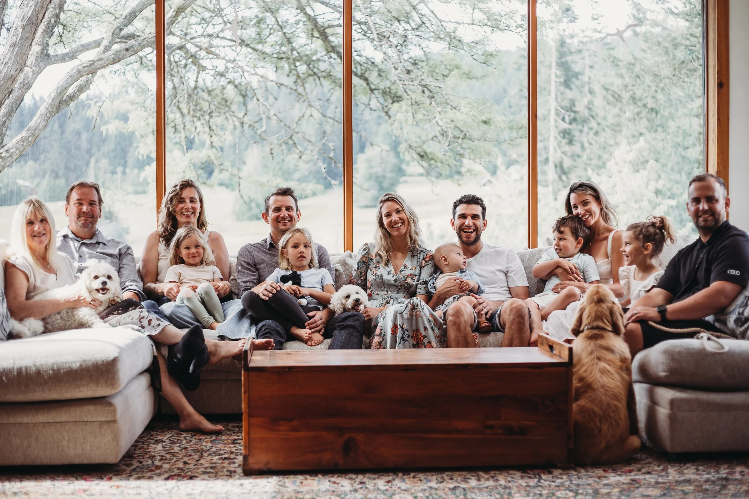 A large family sitting on a sofa in a bright living room with large windows overlooking greenery, featuring several adults, children, and two dogs, all smiling.