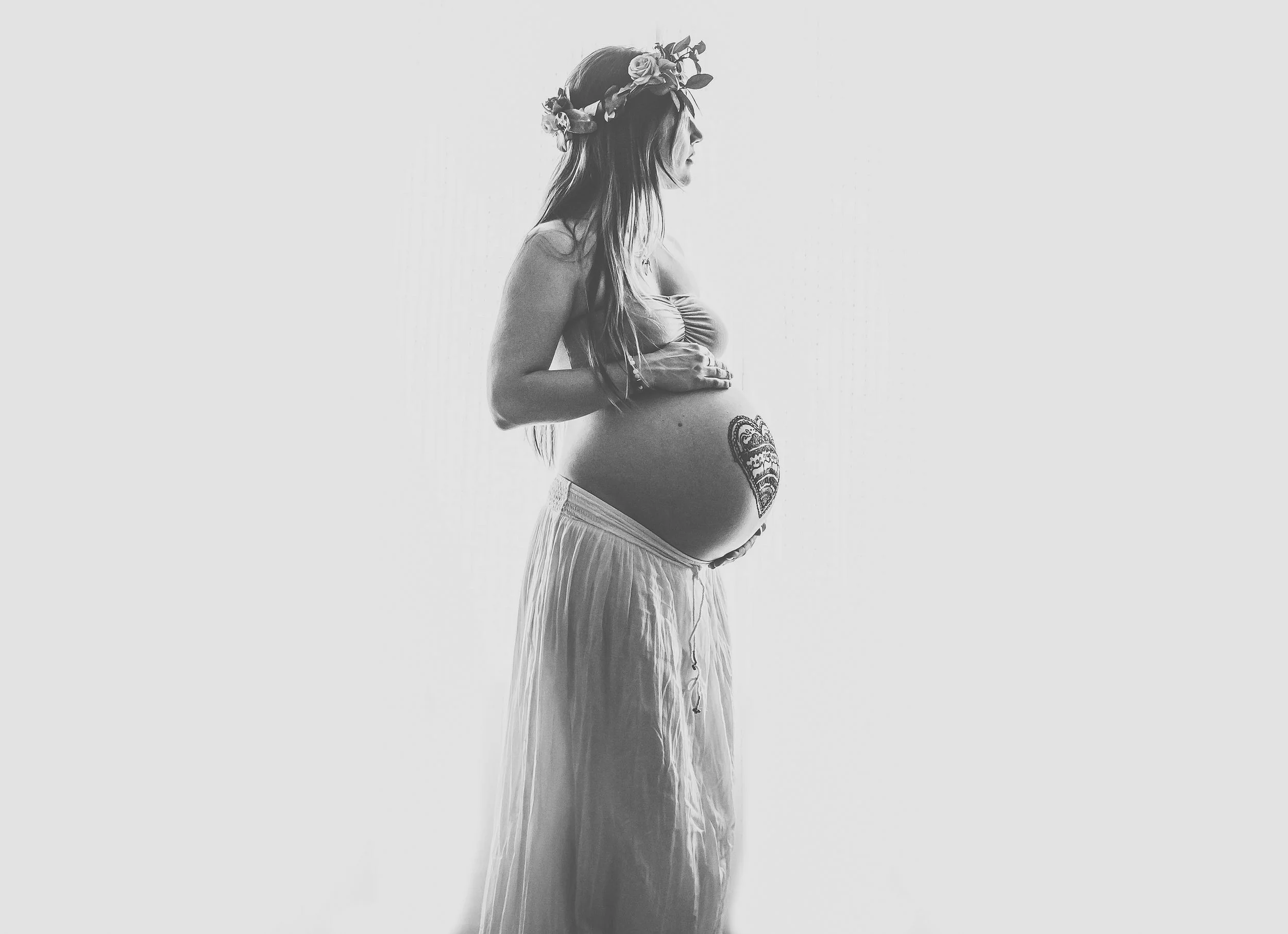 Black and white silhouette of a pregnant woman wearing a flower crown and holding her belly with a henna heart design.