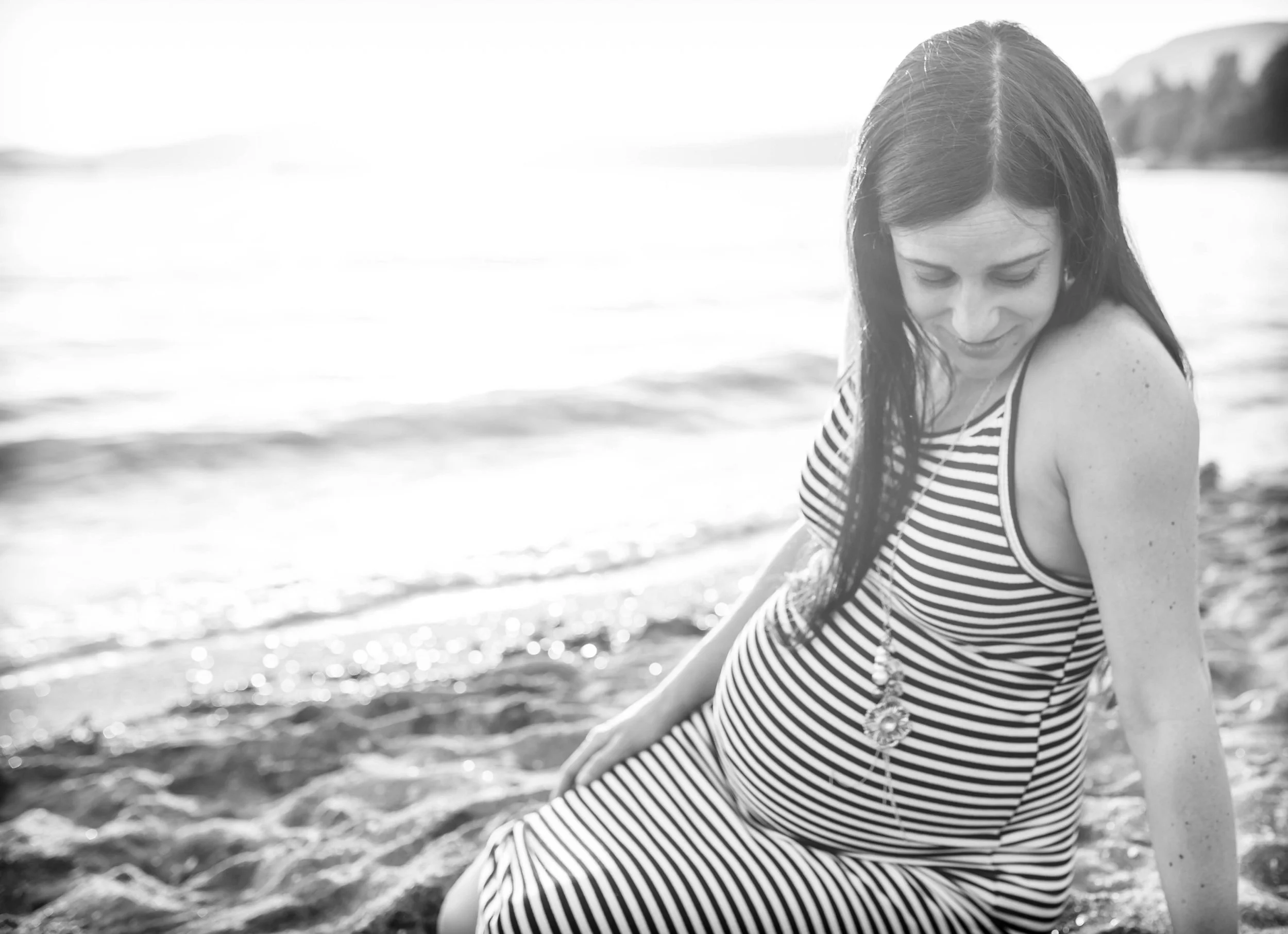 Pregnant woman in striped dress sitting on the beach, smiling and looking down.