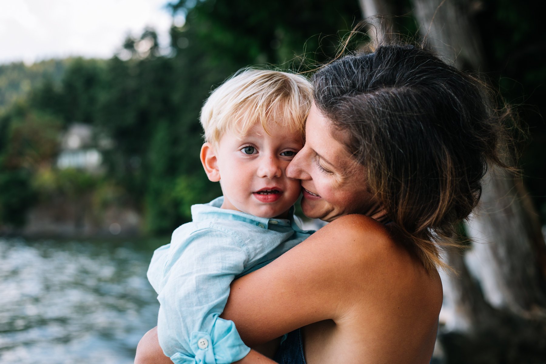 Woman holding young child near a lake with a forest background.