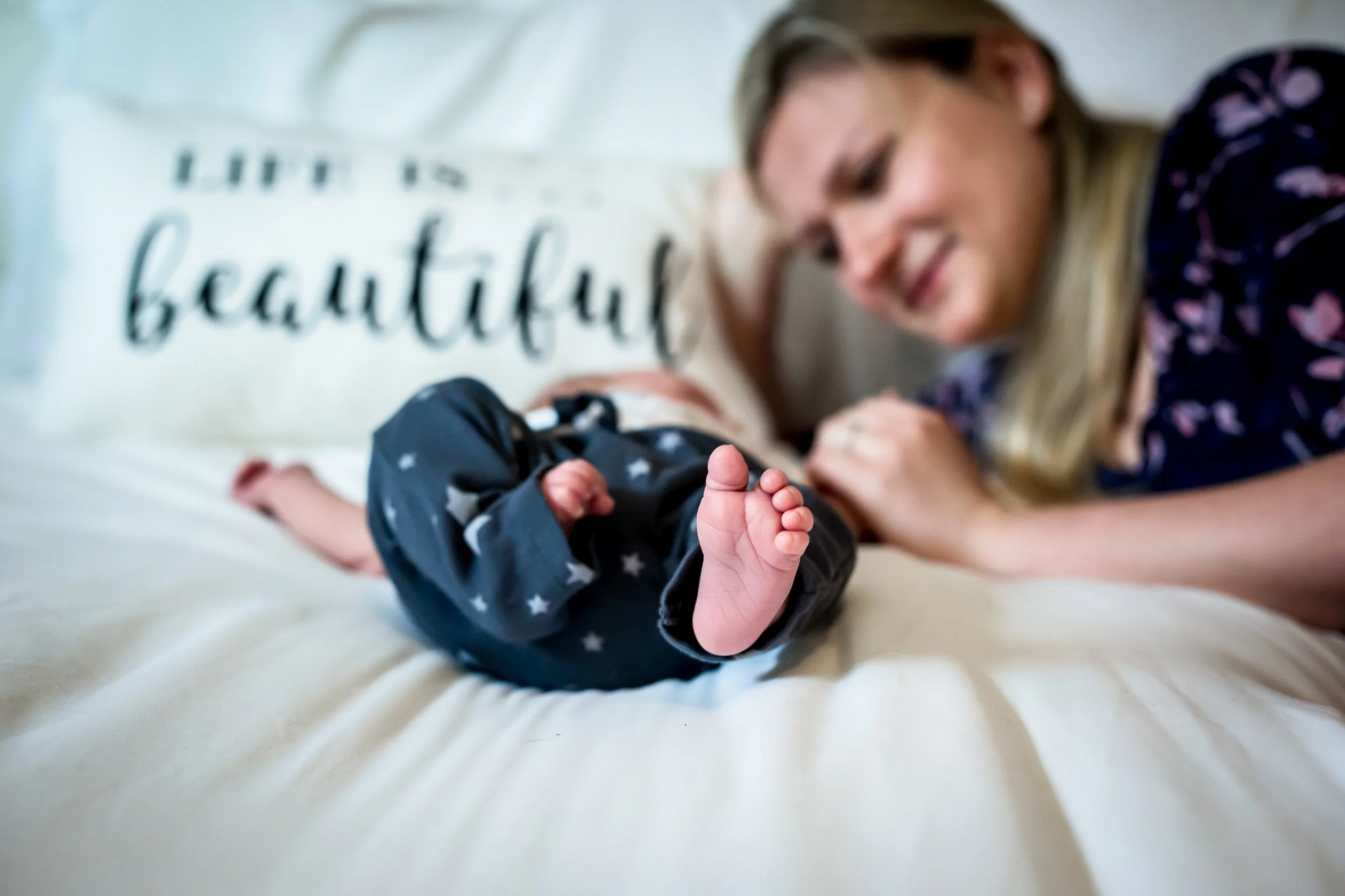 Mother smiling at newborn baby lying on bed with "life is beautiful" pillow in background.