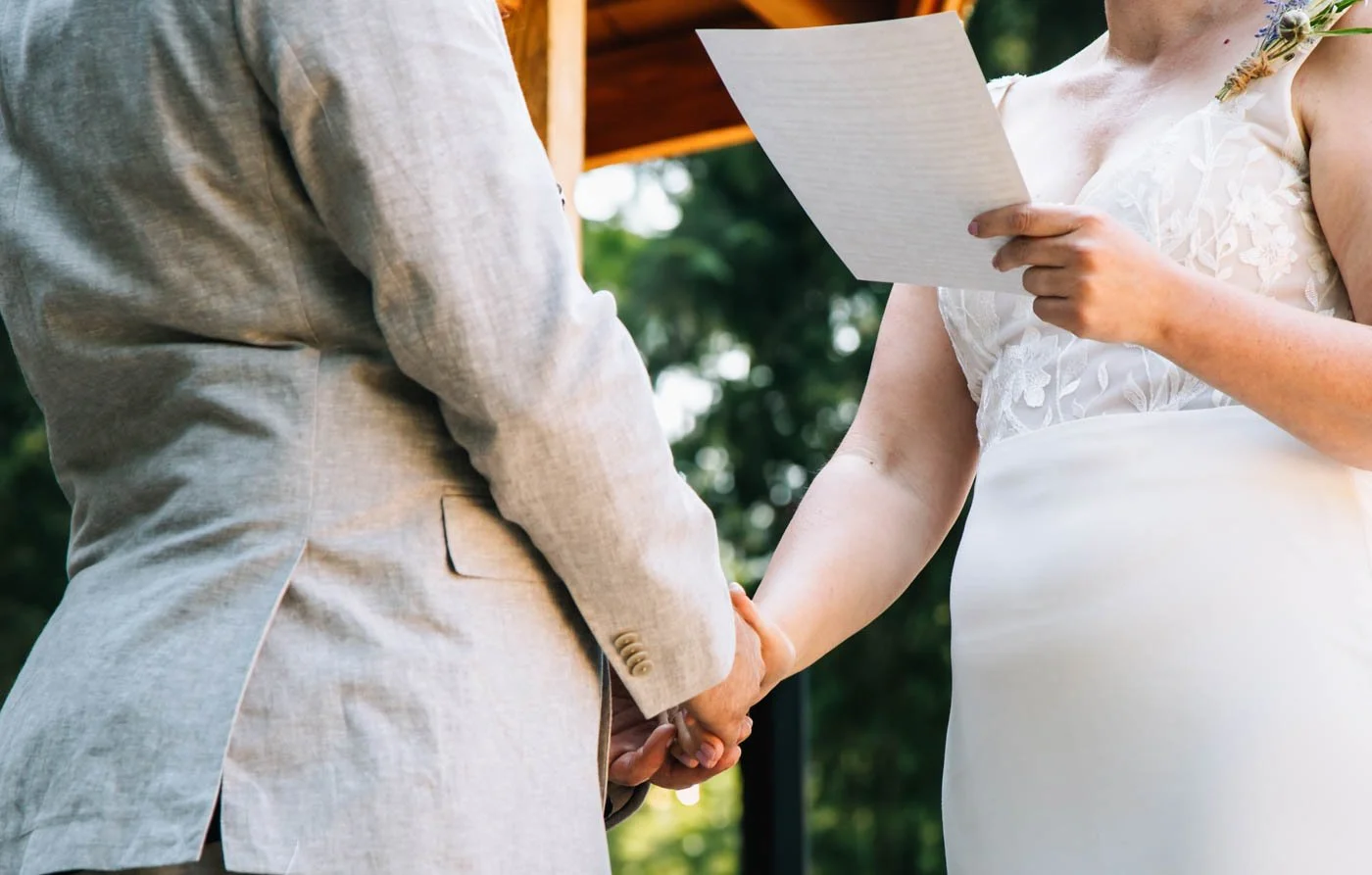 Bride and groom holding hands during wedding ceremony, with the bride holding a paper in one hand.