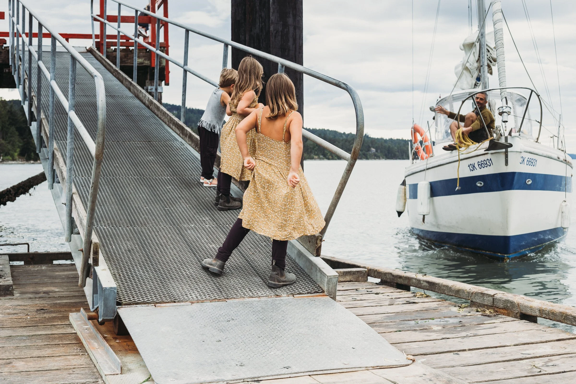 Children on a dock ramp watching a man on a sailboat.