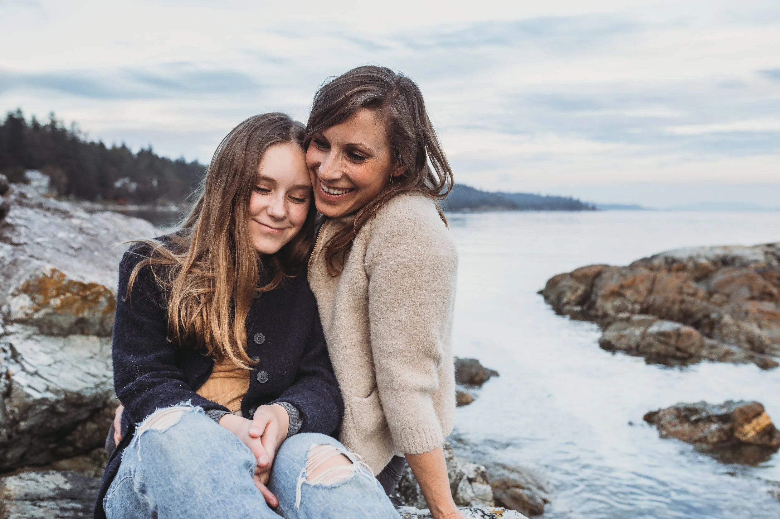 Mother and daughter hugging by the seaside, rocky shore in the background.