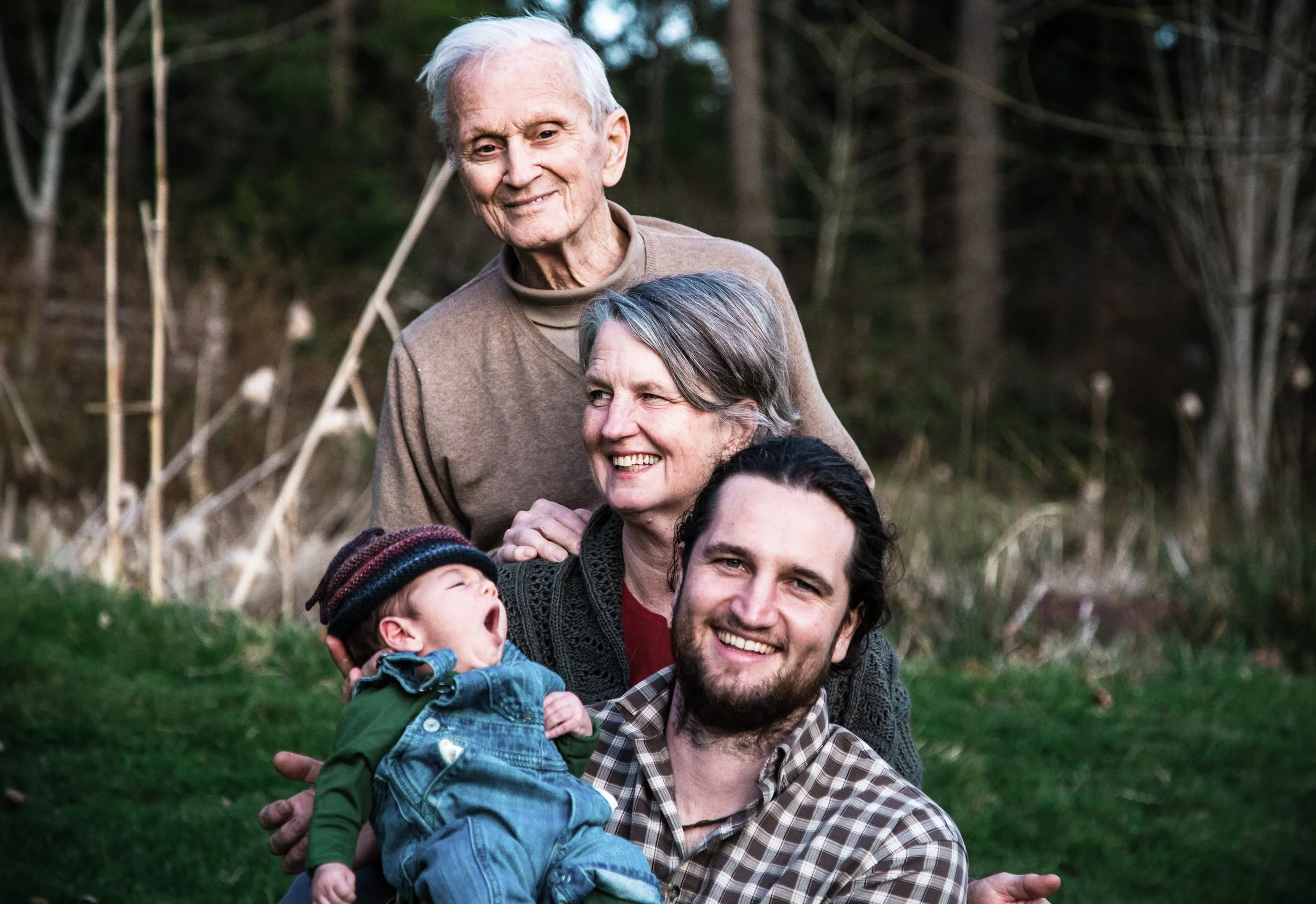 Four people outdoors, including an elderly man, a woman, a man with a beard, and a yawning baby wearing a cap and denim overalls, in a natural, grassy setting.
