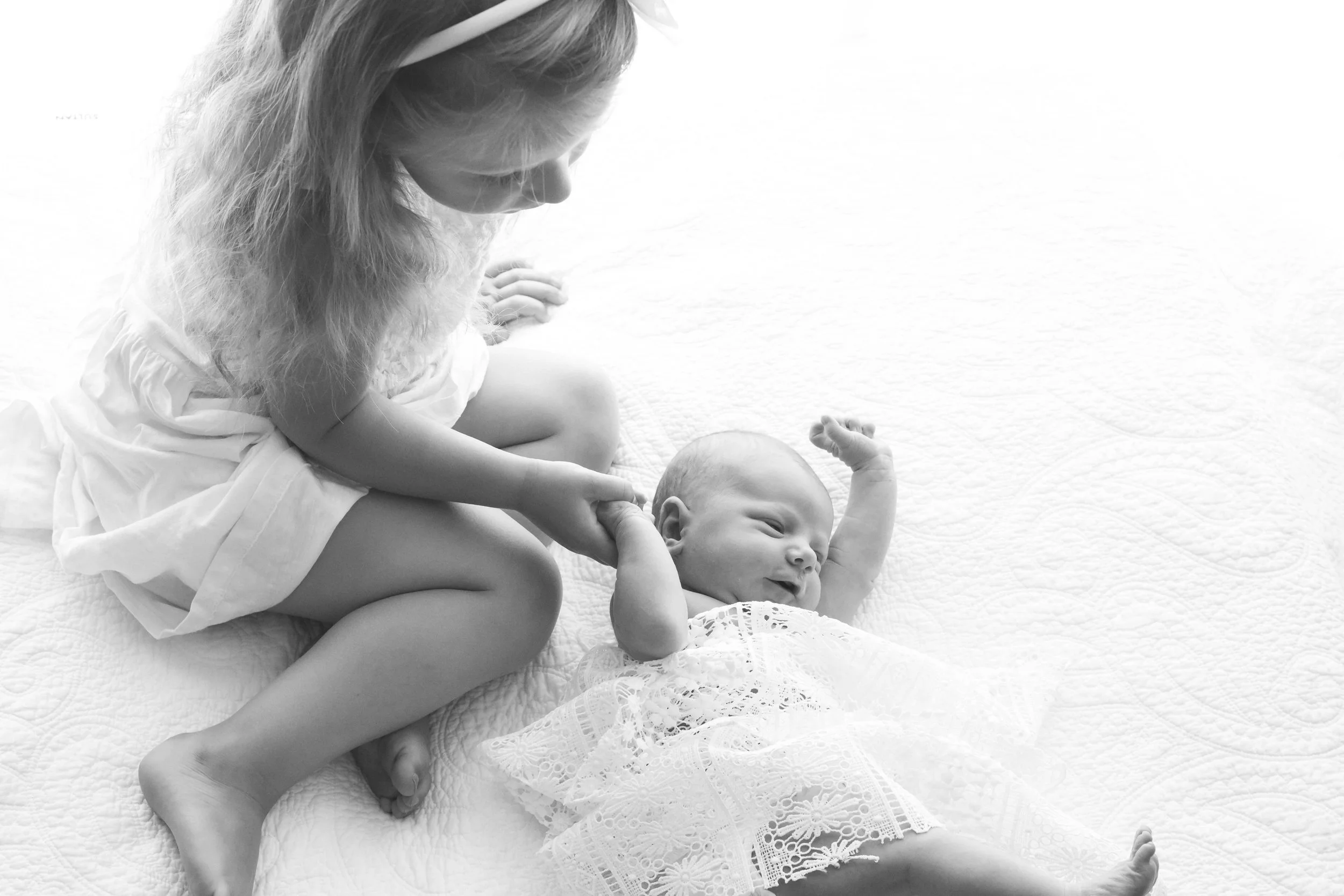 Black and white photo of a young girl sitting beside a baby dressed in lace, both on a quilted surface.