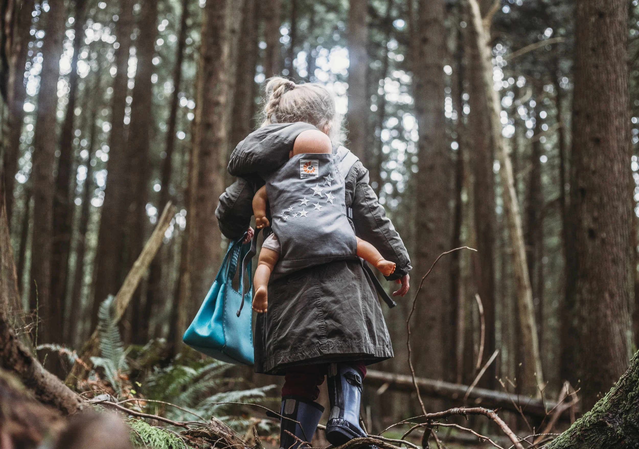 Child walking in forest with doll in backpack carrier, carrying blue bag.