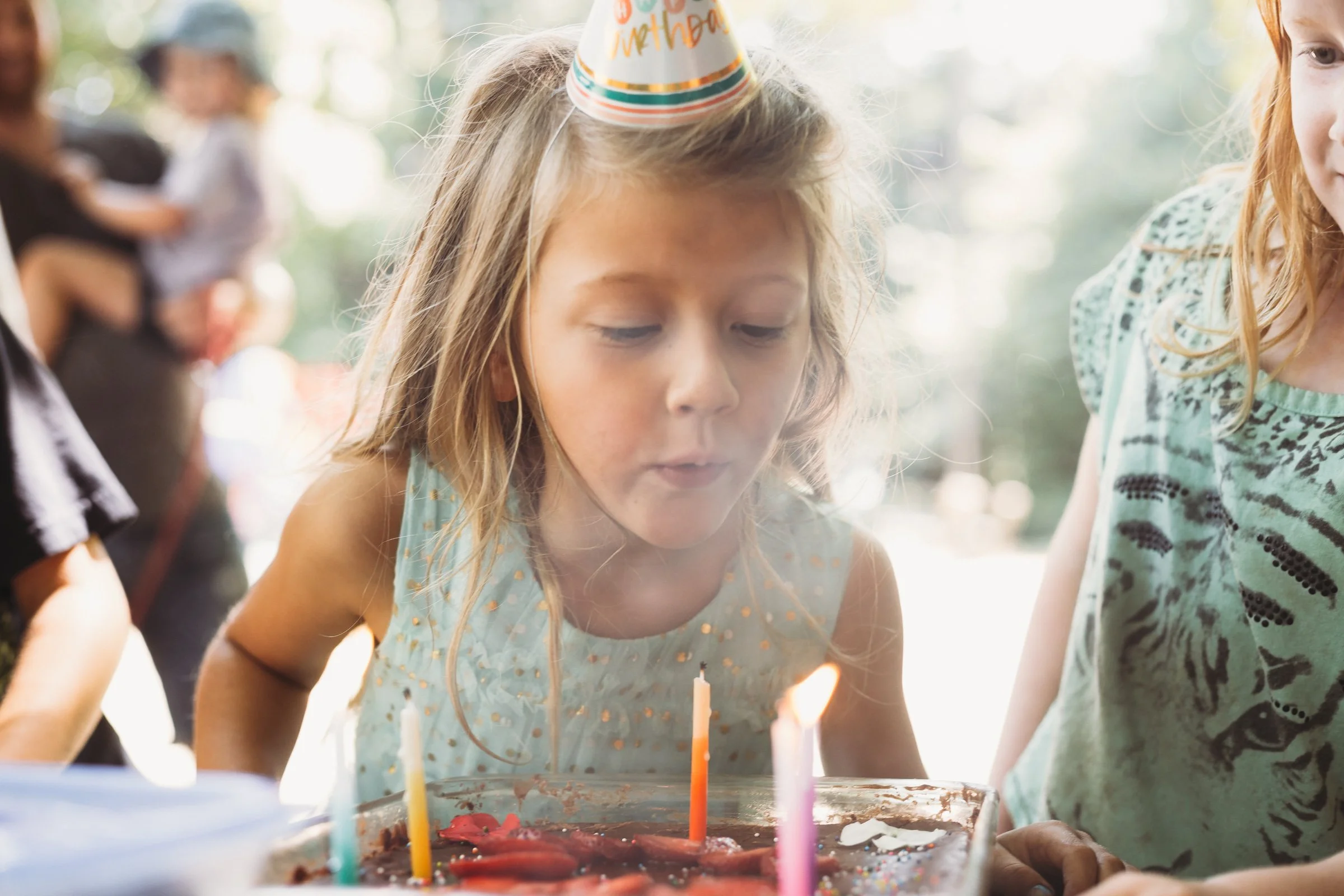Child blowing out birthday candles on a cake, wearing a party hat, surrounded by friends.