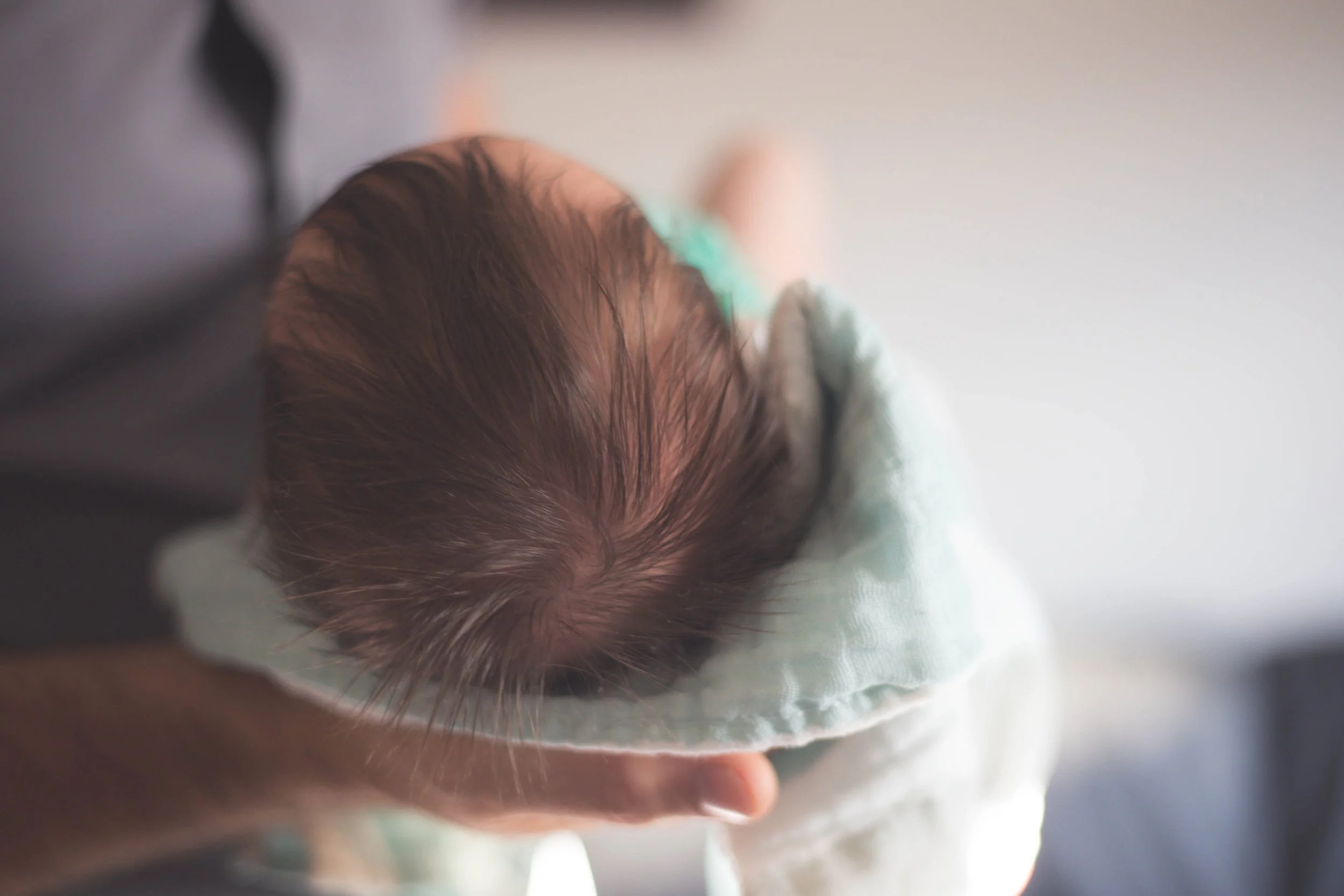 Baby with dark hair cradled in light blanket, viewed from the back.