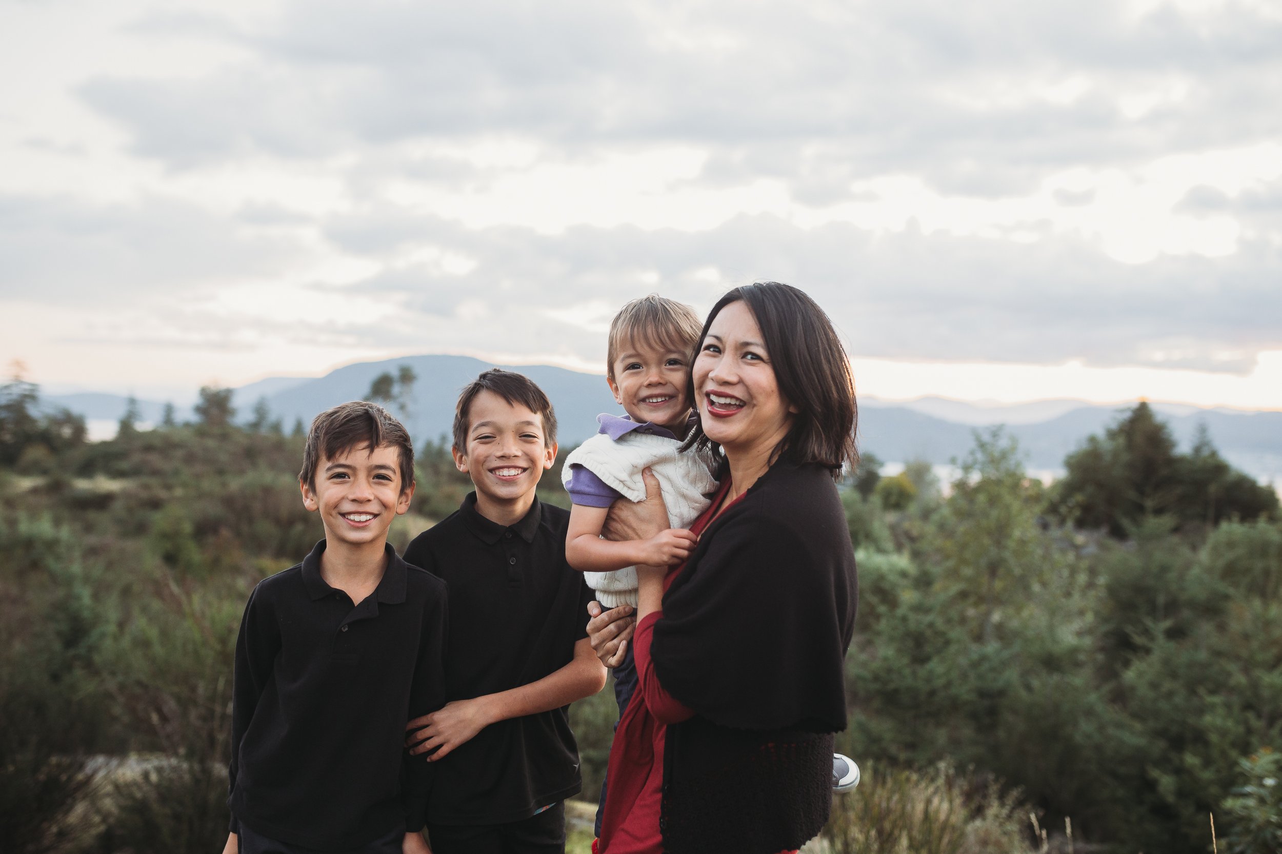 A woman and three children smiling outdoors with a scenic mountainous backdrop.