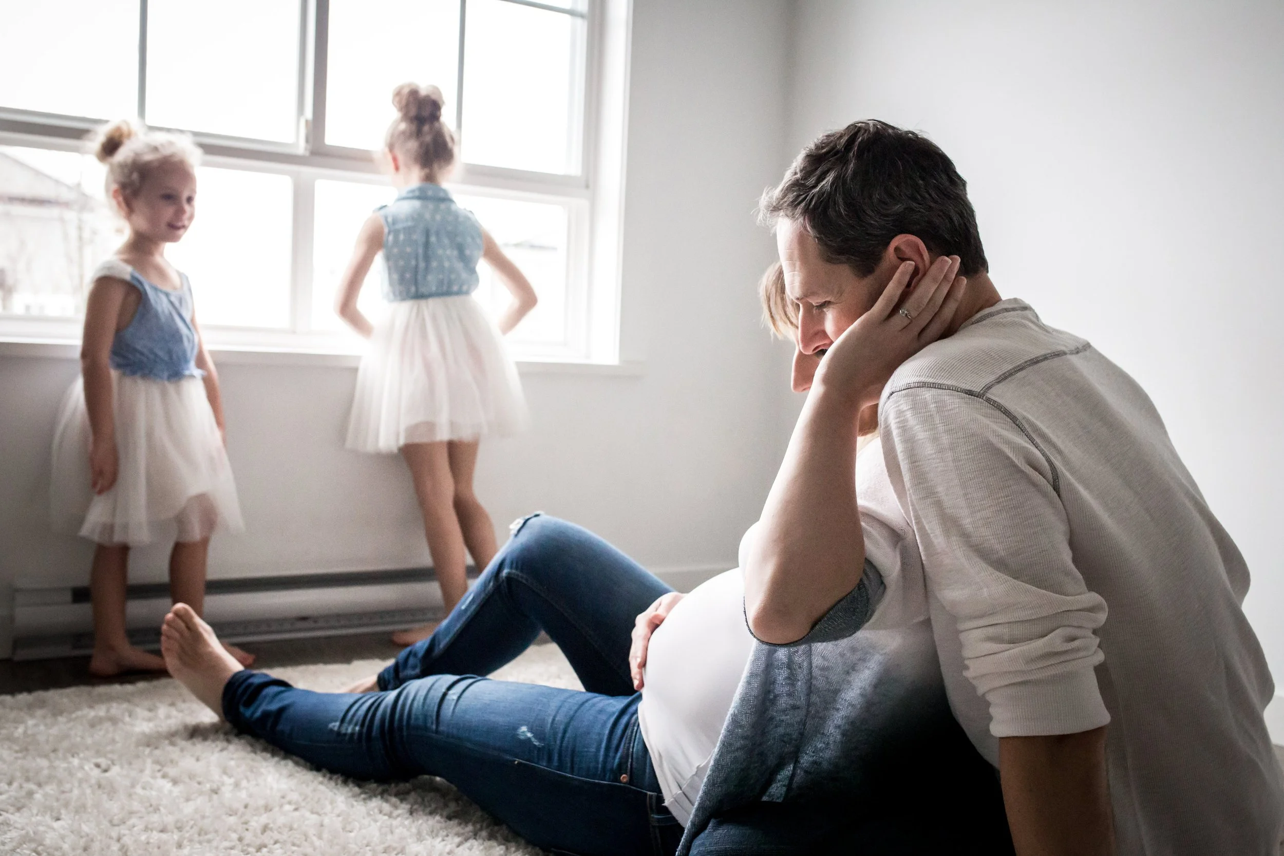 Family with children at home, pregnant woman, man embracing, two girls by window, bright room, casual atmosphere.