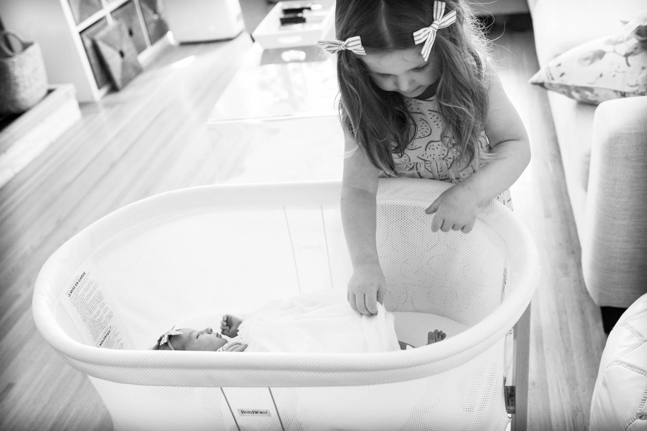 Young girl looking at baby in bassinet, home setting, black and white photo.