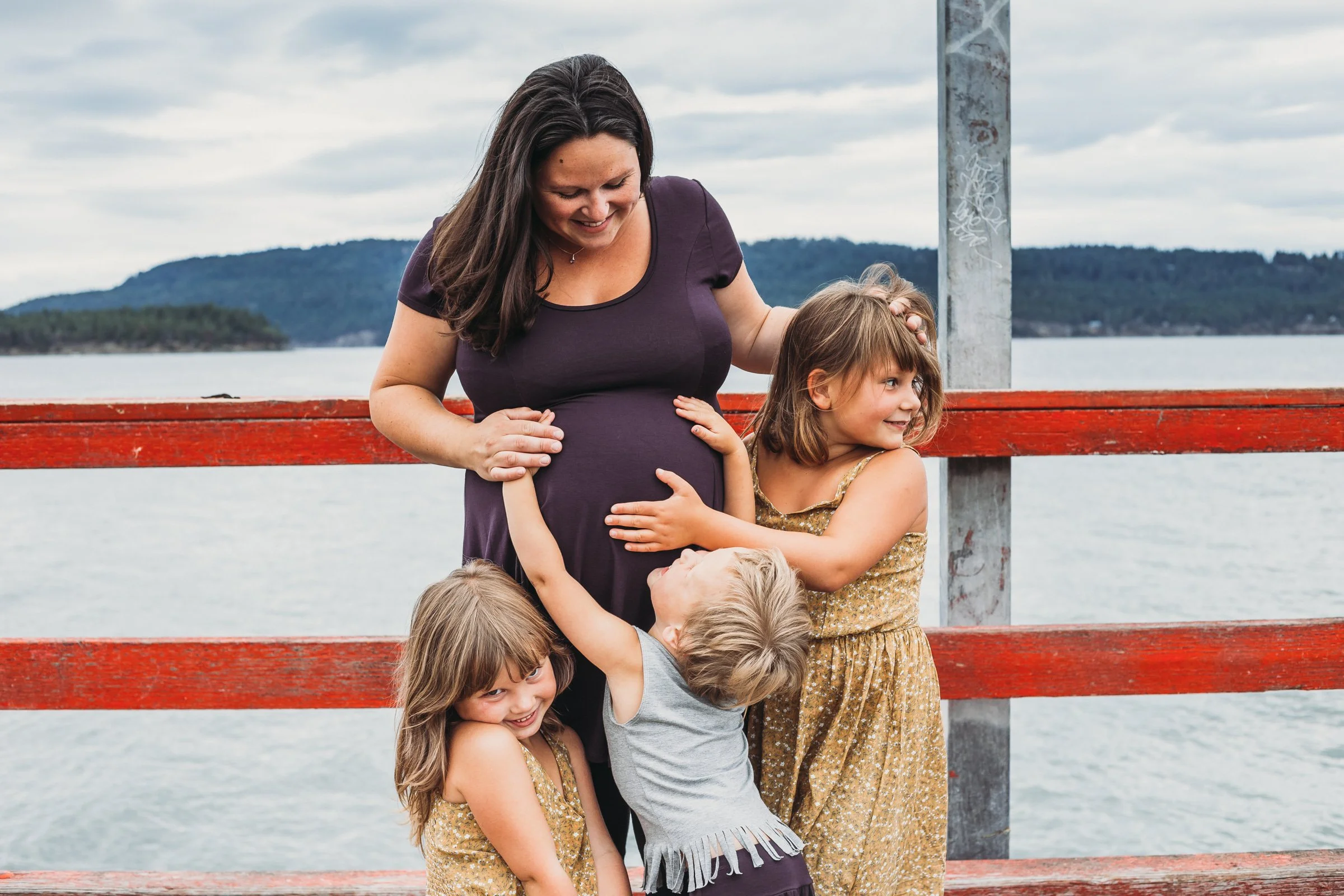 A pregnant woman smiling with three children around her, outdoors by a body of water and a red railing.