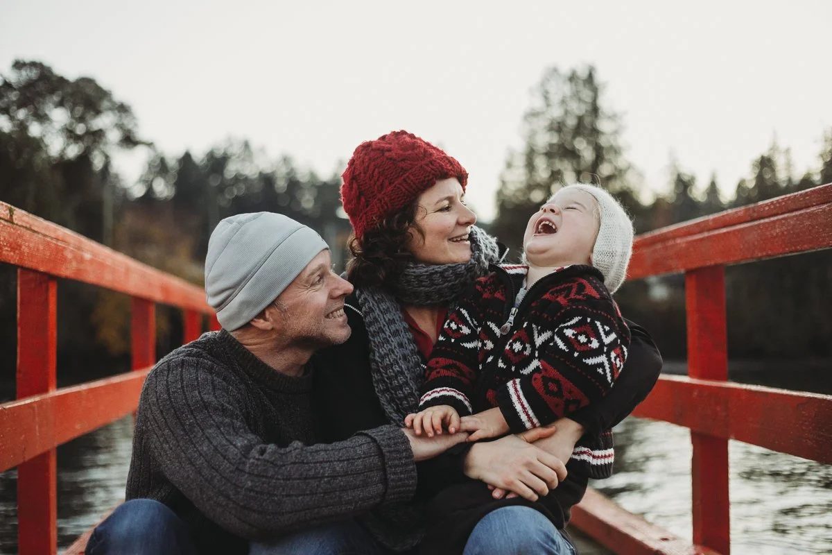 Family sitting on bridge, smiling and laughing, wearing winter clothing.