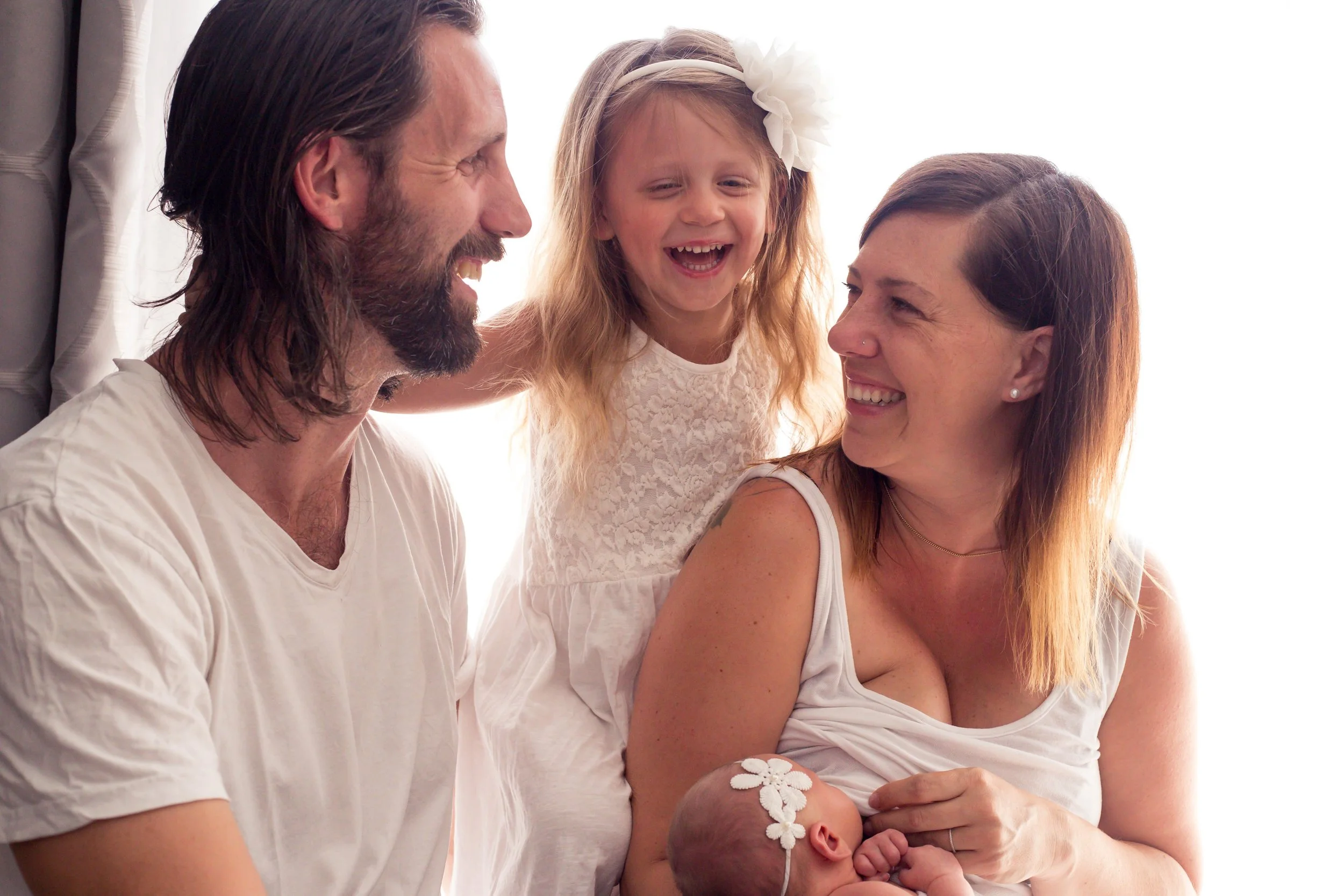 Happy family with mother, father, young daughter, and newborn baby in white clothing, smiling and sitting together in a bright room.