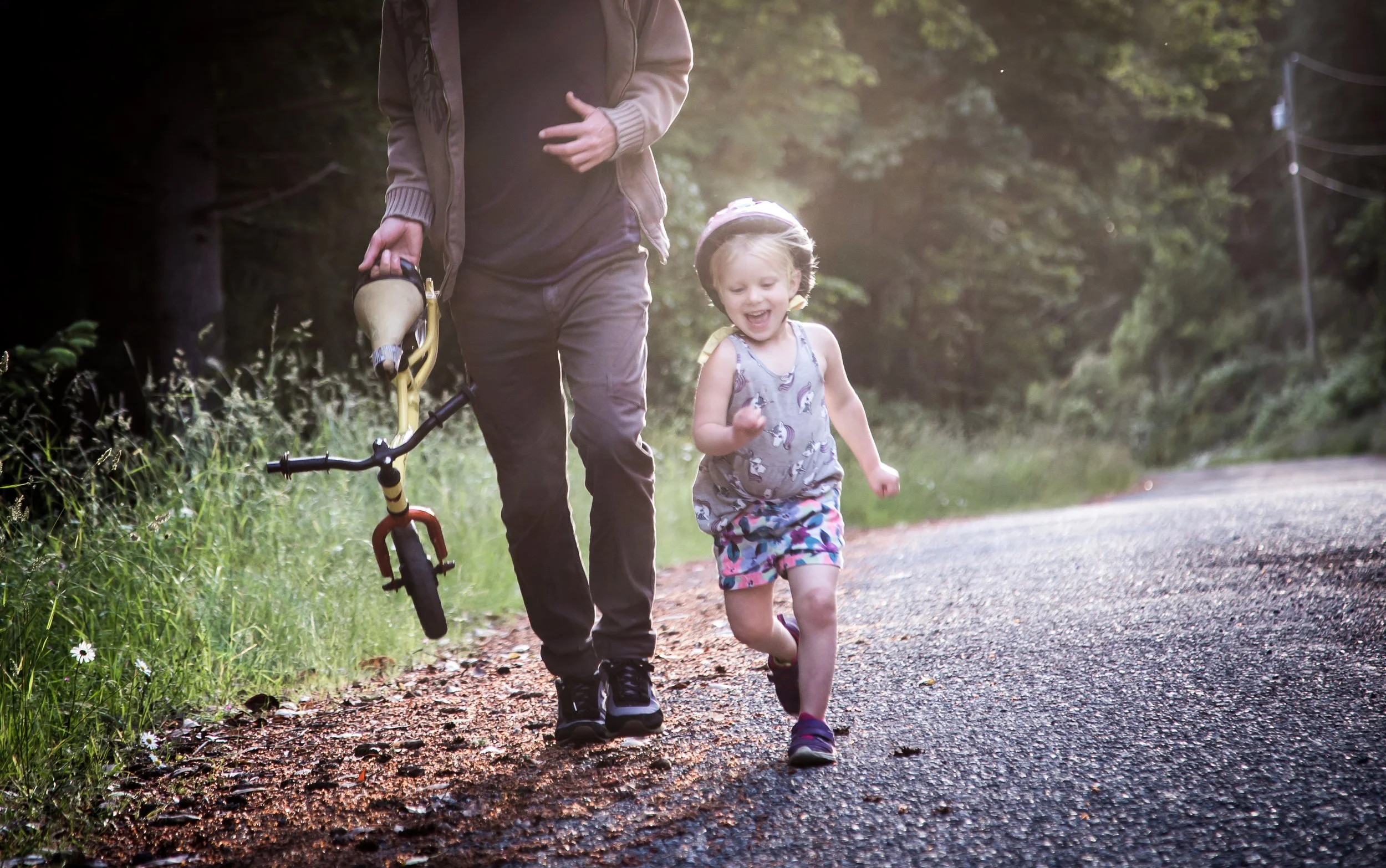 A child running along a path next to an adult holding a bicycle, surrounded by greenery and sunlight.