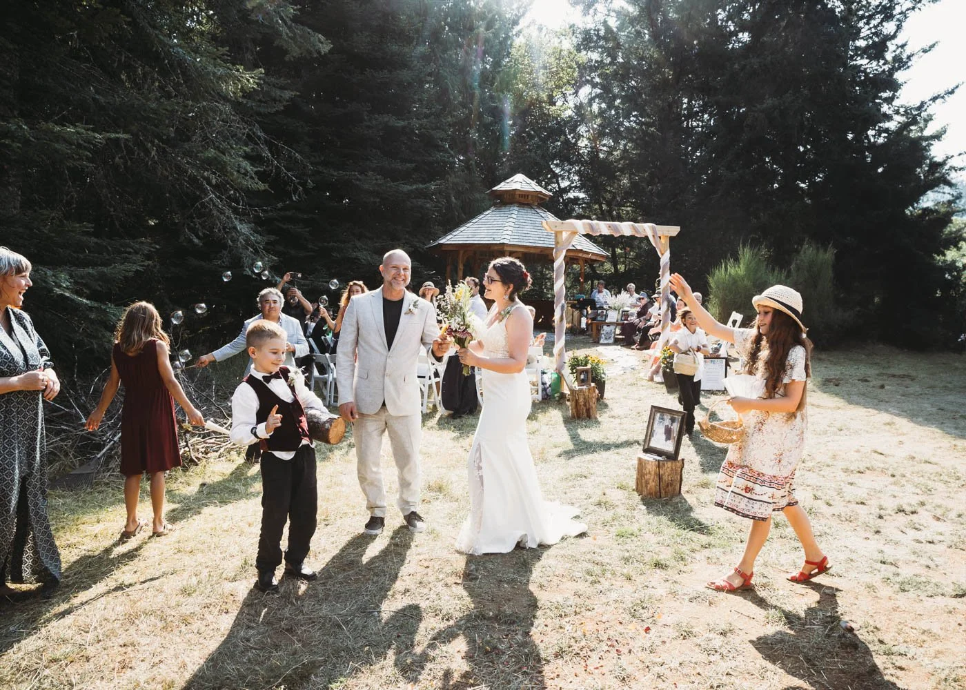 Outdoor wedding ceremony with bride and guests, featuring a rustic wooden structure and forest backdrop.