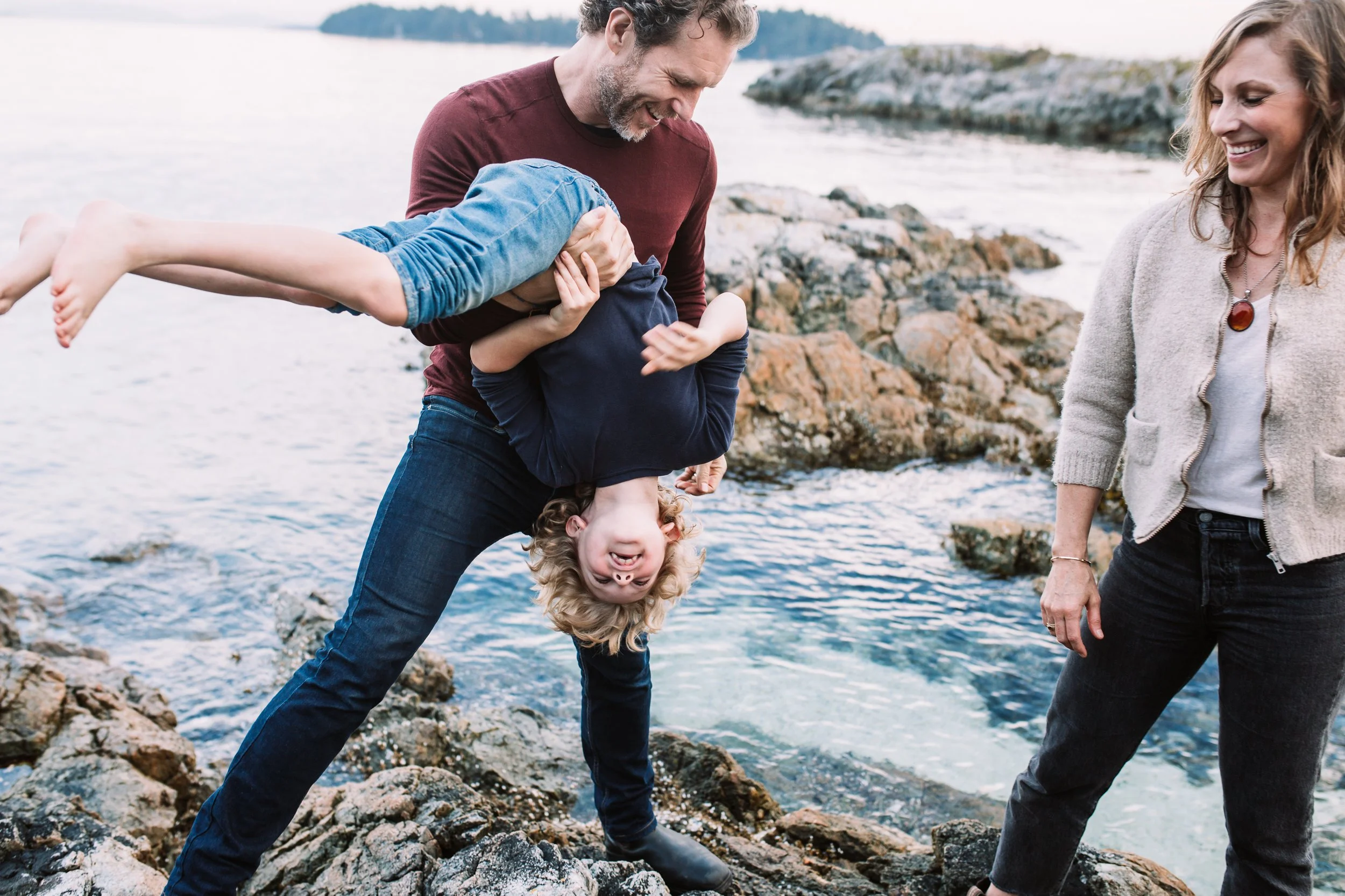 Family playing by rocky seaside, man holding child upside down, woman smiling.