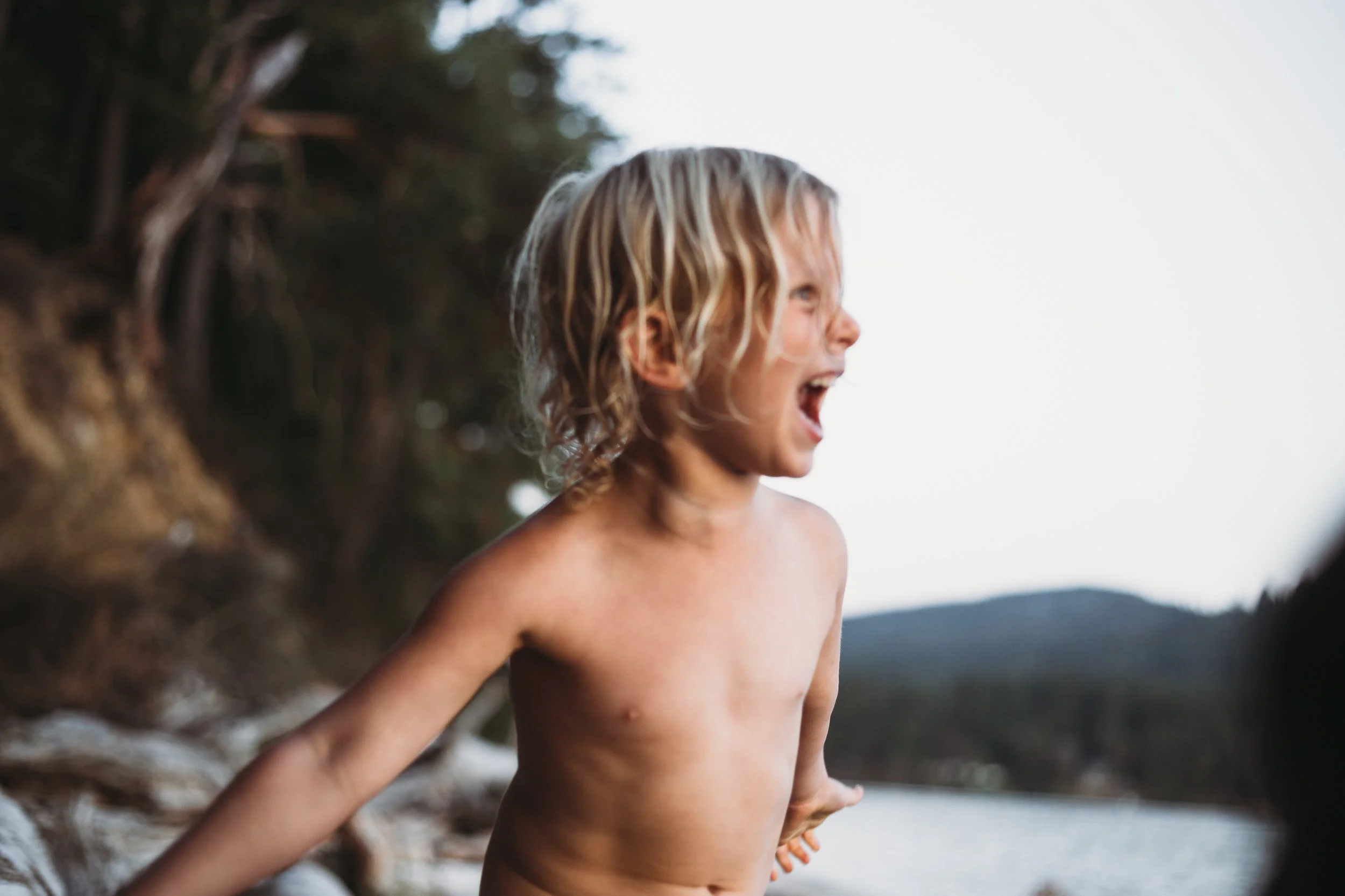 Child with blond hair laughing outdoors near a forest and a body of water.