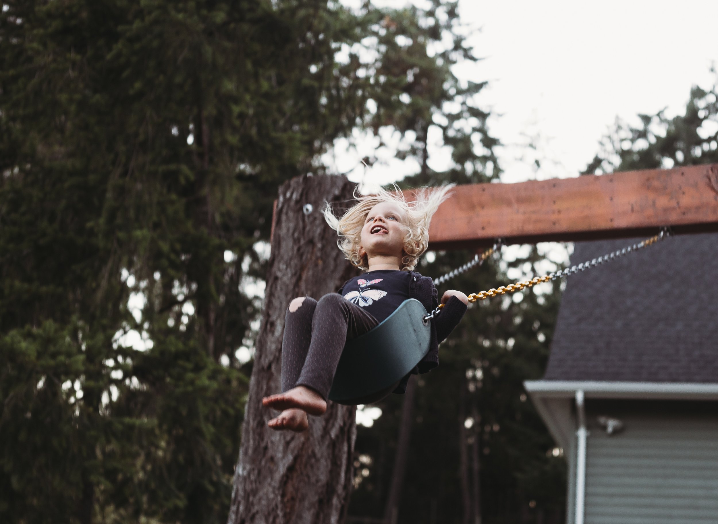 Child swinging joyfully on a swing set in a backyard with trees in the background.