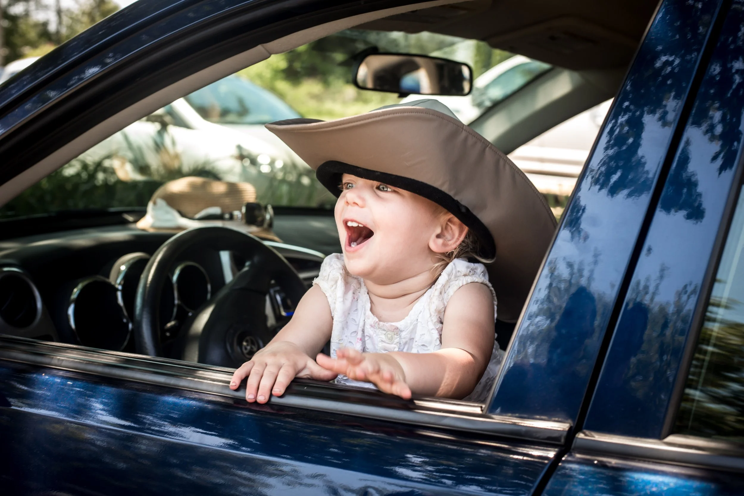 Happy child wearing a large hat leaning out of a car window, smiling and laughing.