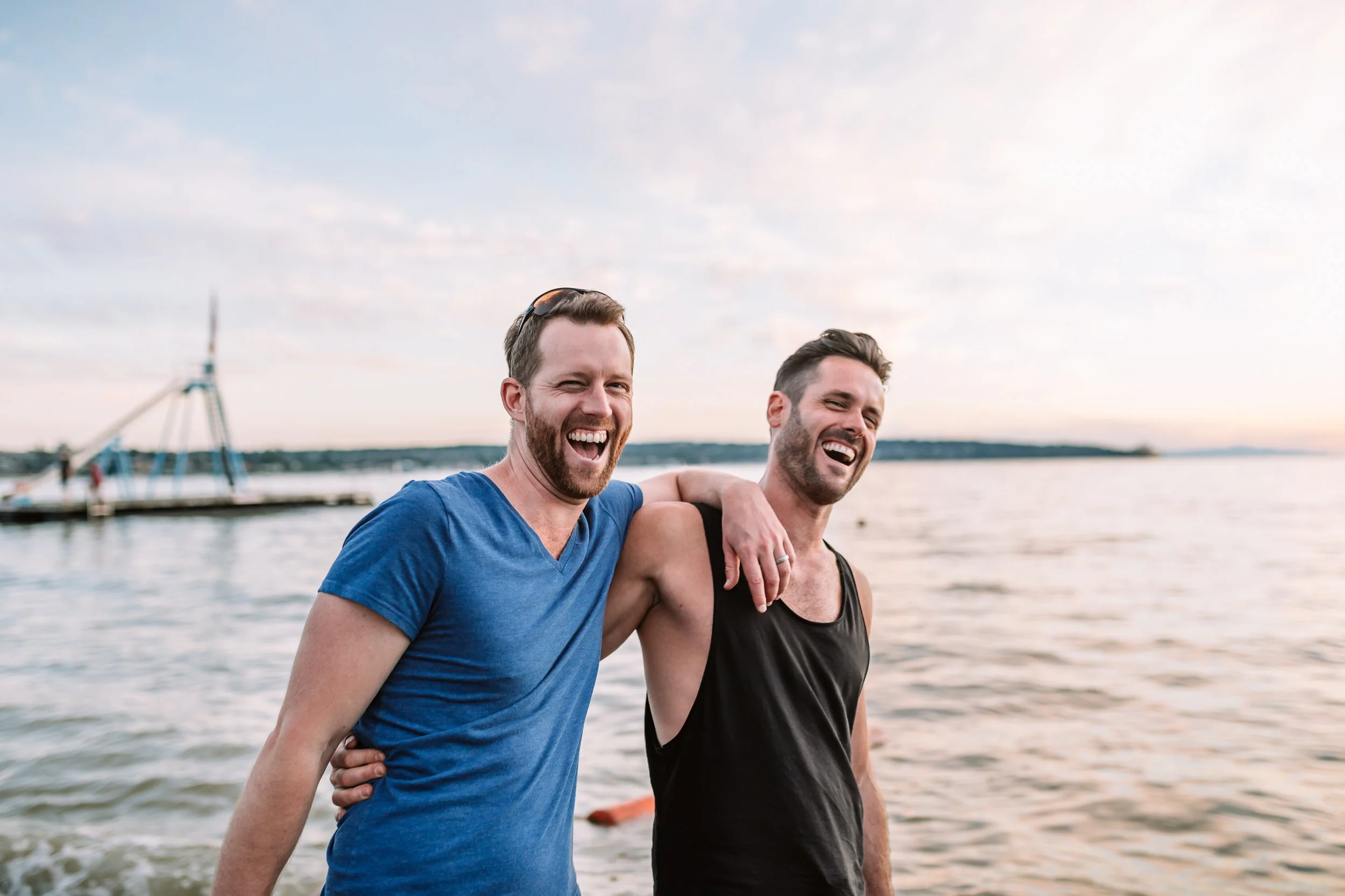 Two men laughing, standing by the water at sunset.