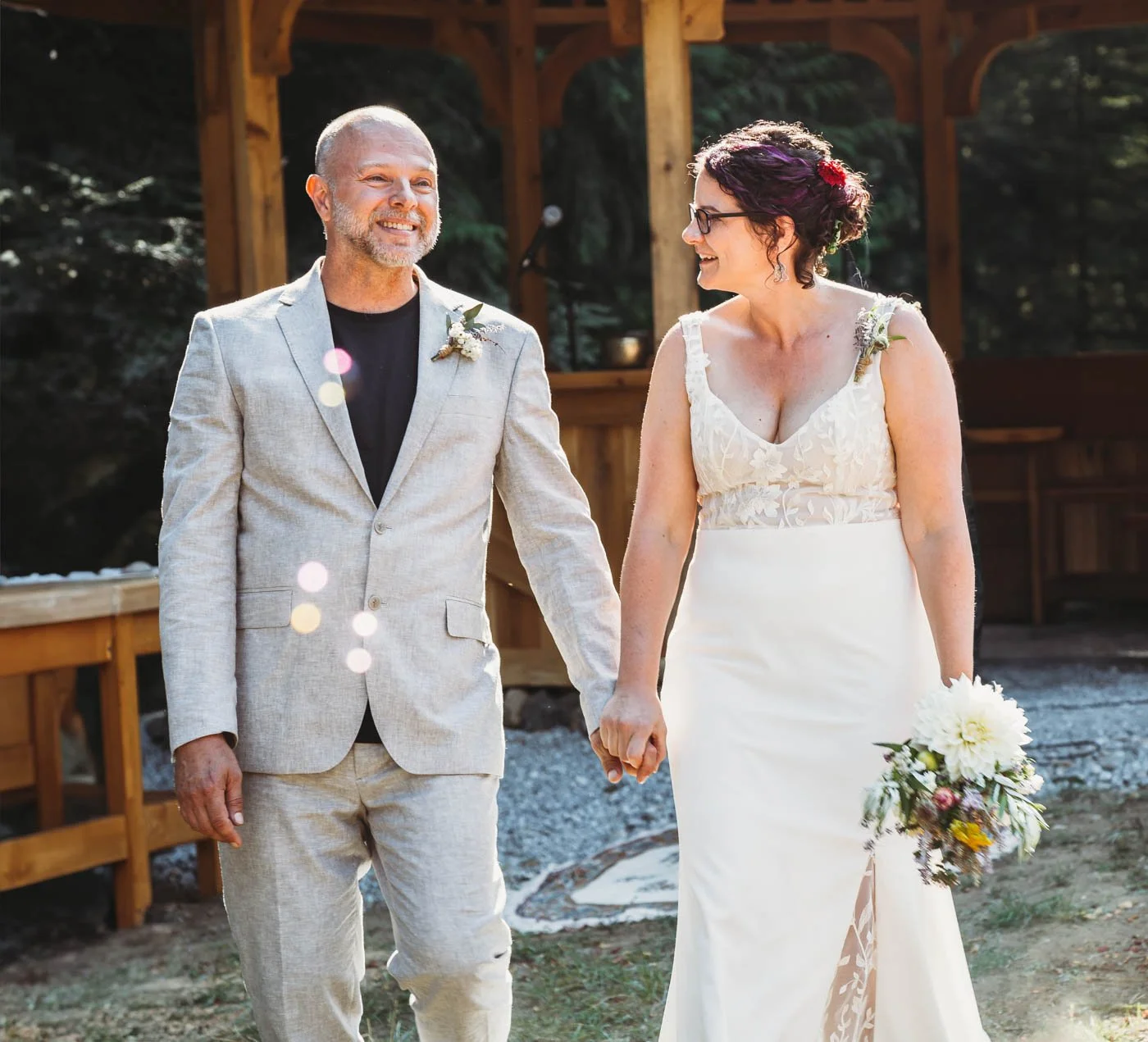 A couple holding hands at an outdoor wedding ceremony, with the man wearing a light-colored suit and the woman in a white wedding dress, carrying a bouquet.