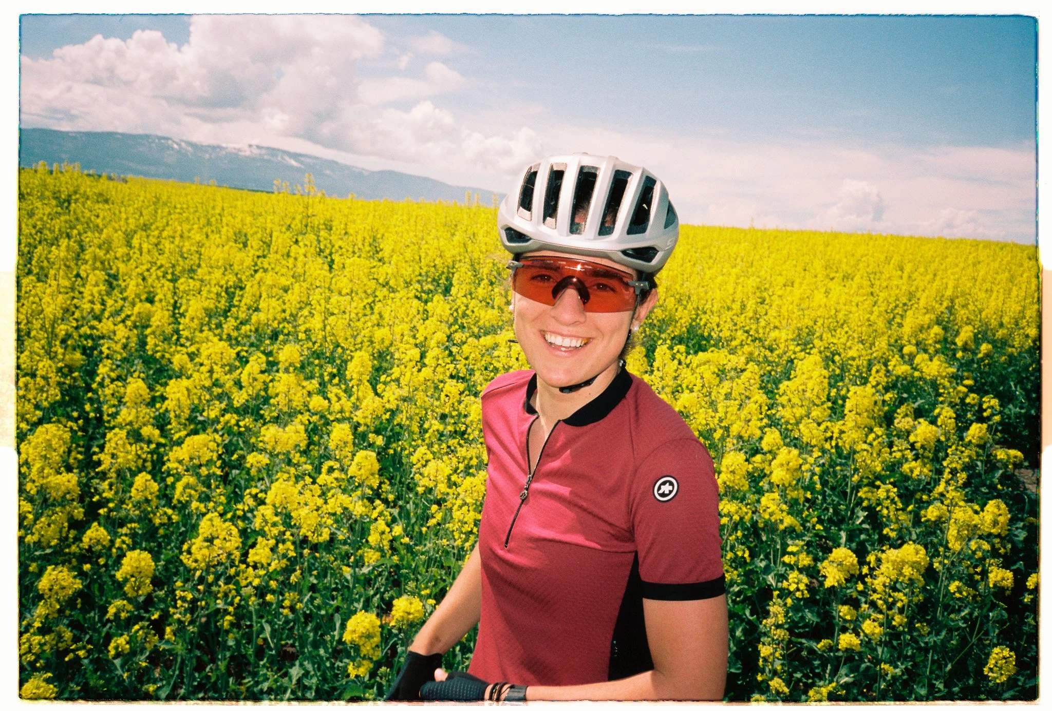 Woman in bicycling helmet, sunglasses and red Assos jersey smiles at the camera in front of a field of yellow canola flowers