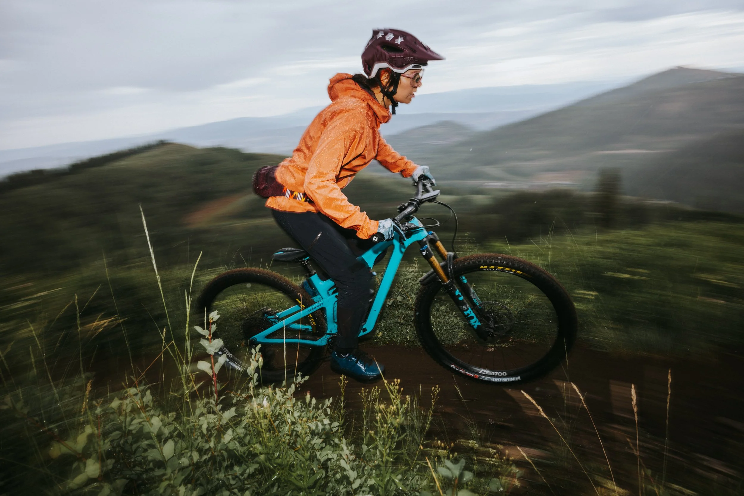 Woman on blue Yeti mountain bike, wearing orange Patagonia rain jacket, maroon Fox helmet, behind slightly burred foreground of plants and grass with gray skies and mountain backdrop