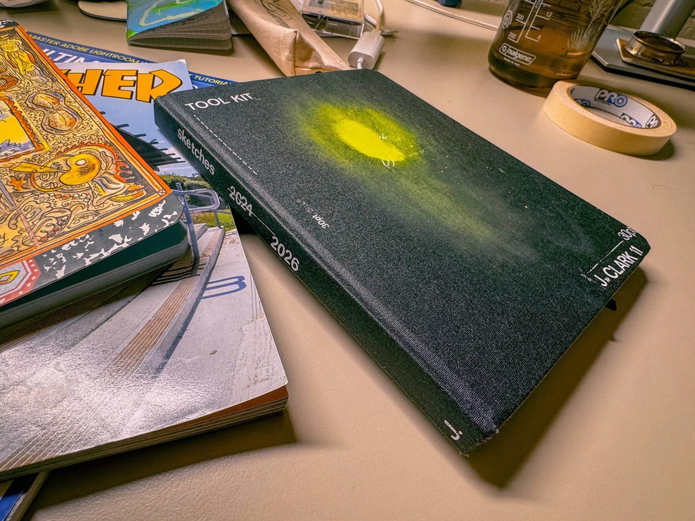 A black sketch book with white lettering, sketches 2024-2026, Tool Kit, J Clark 11, on top of a Thrasher Skateboarding magazine on a desk, closely cropped