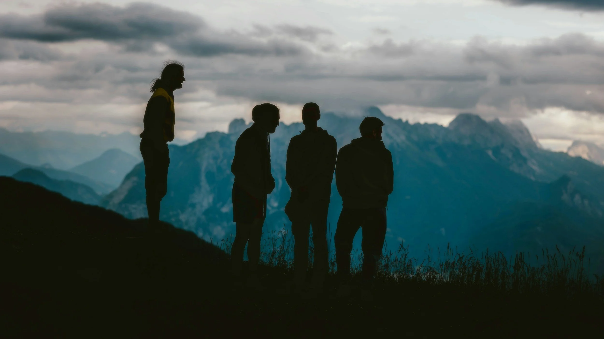 Silhouettes of four people standing on a hillside, overlooking a mountain range and cloudy sky.