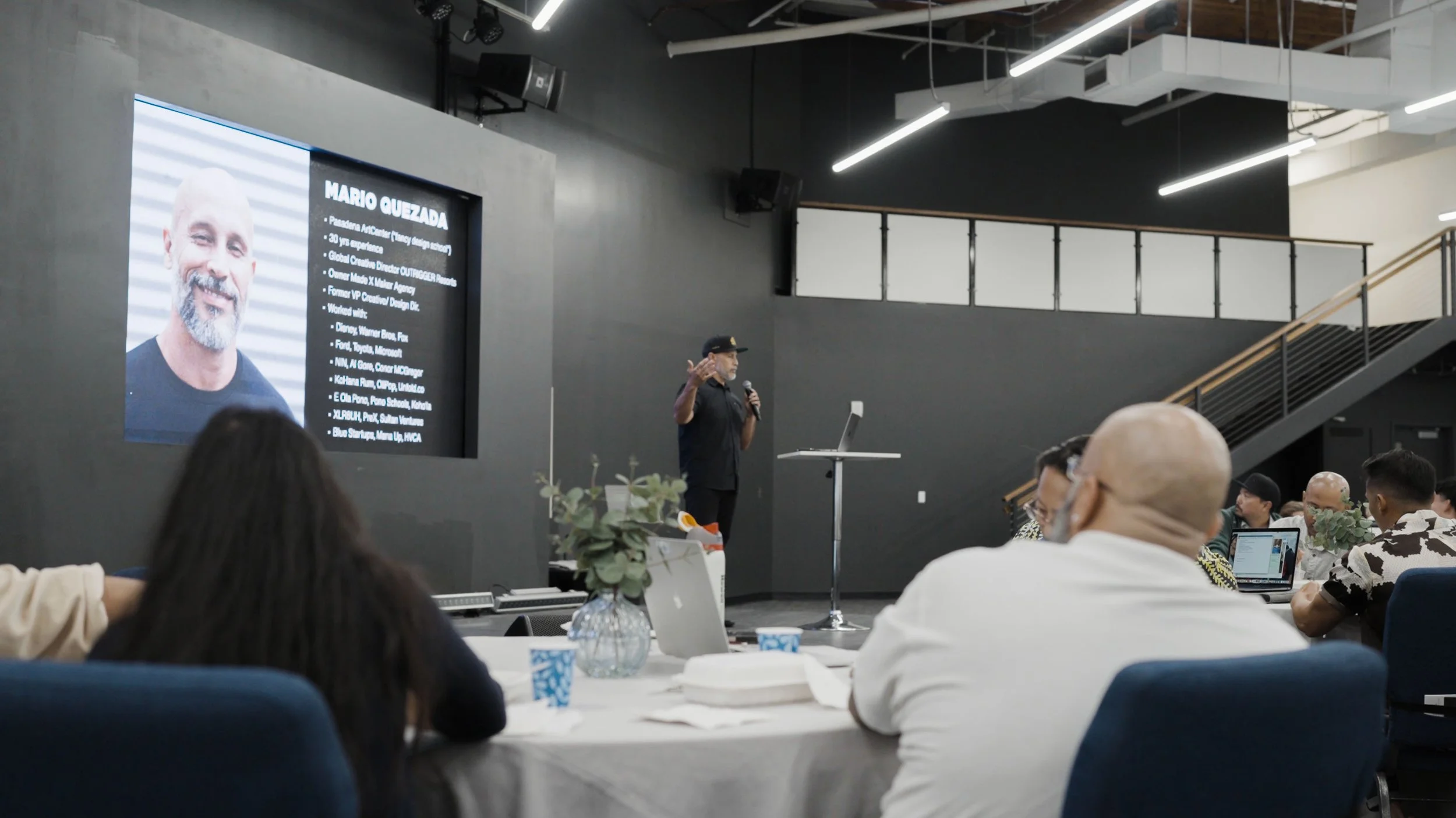 A speaker on stage giving a presentation at a conference with a large screen displaying a photo and biography of Mario Guezada, visible in the background. Audience members are seated at tables, some taking notes.