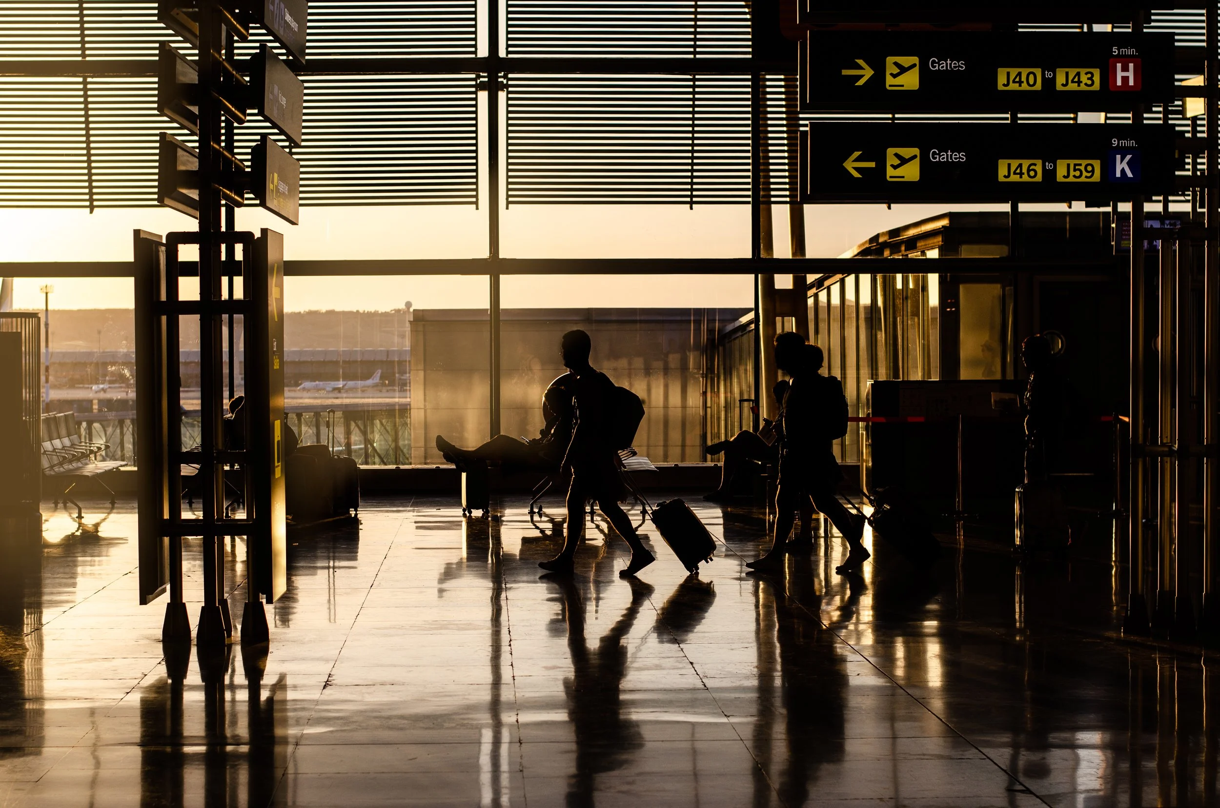 Silhouettes of travelers walking with luggage inside an airport terminal at sunset, with large windows showing planes outside and departure gate signs hanging overhead.