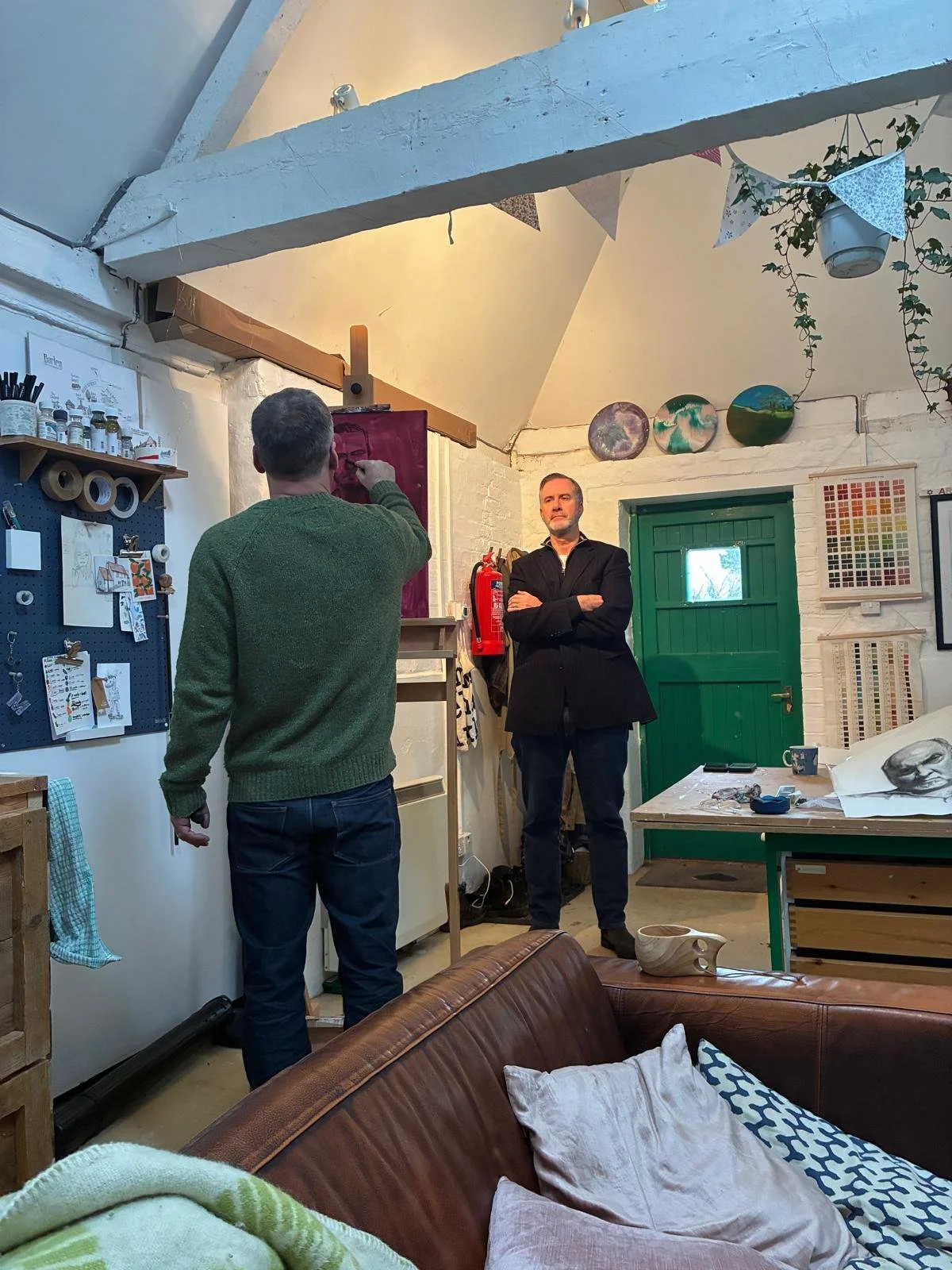 Two men in a room decorated with art and crafts, one painting on an easel and the other standing with arms crossed, while the first man paints or draws on the canvas.