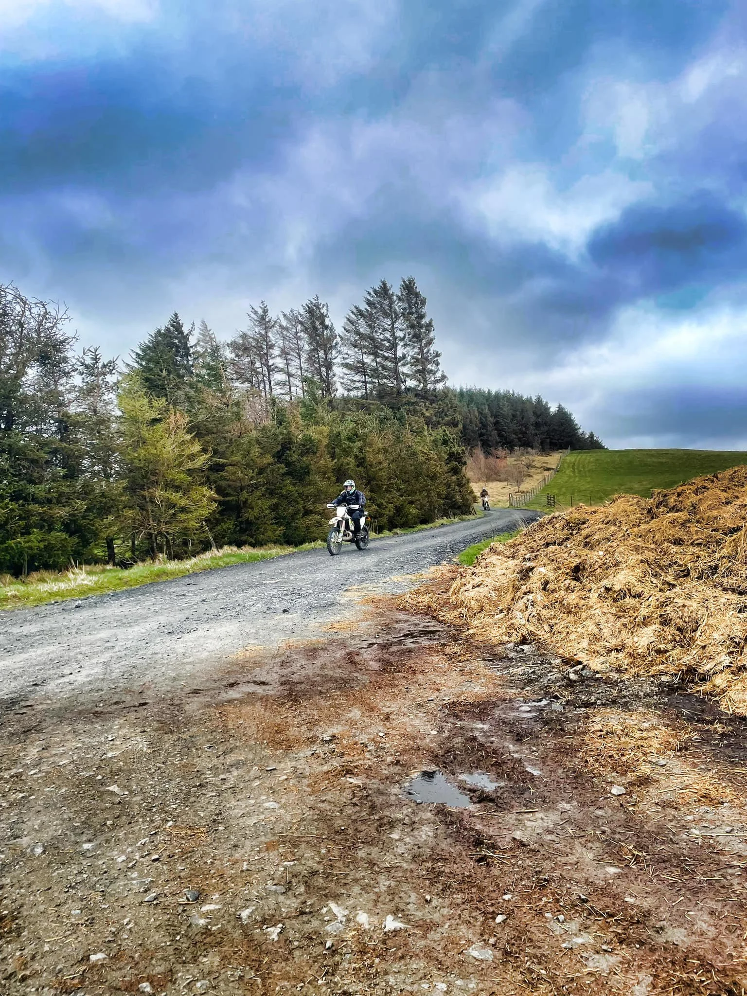 Off-road dirt bike riders on a gravel path through a hilly, wooded area with trees, grass, and cloudy sky.