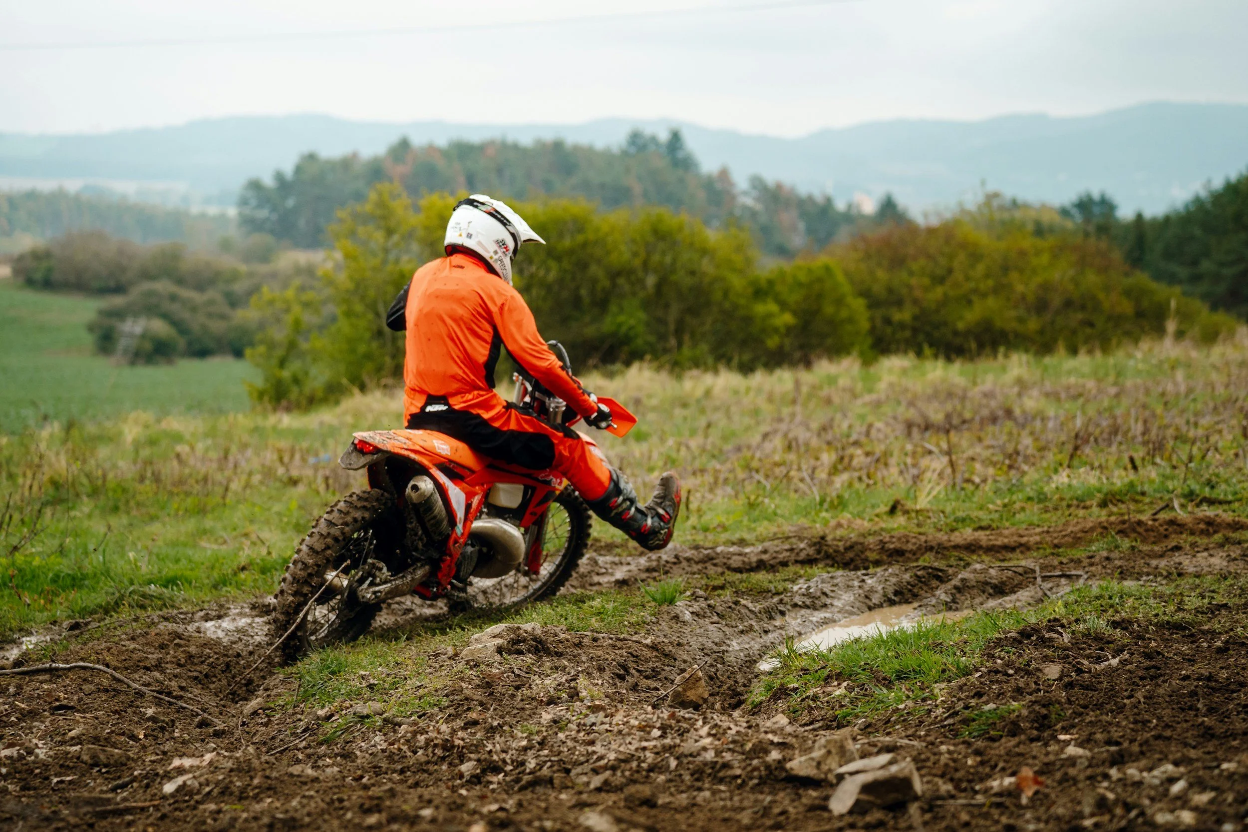A person in orange gear and a white helmet riding a dirt bike on muddy terrain with trees and hills in the background.