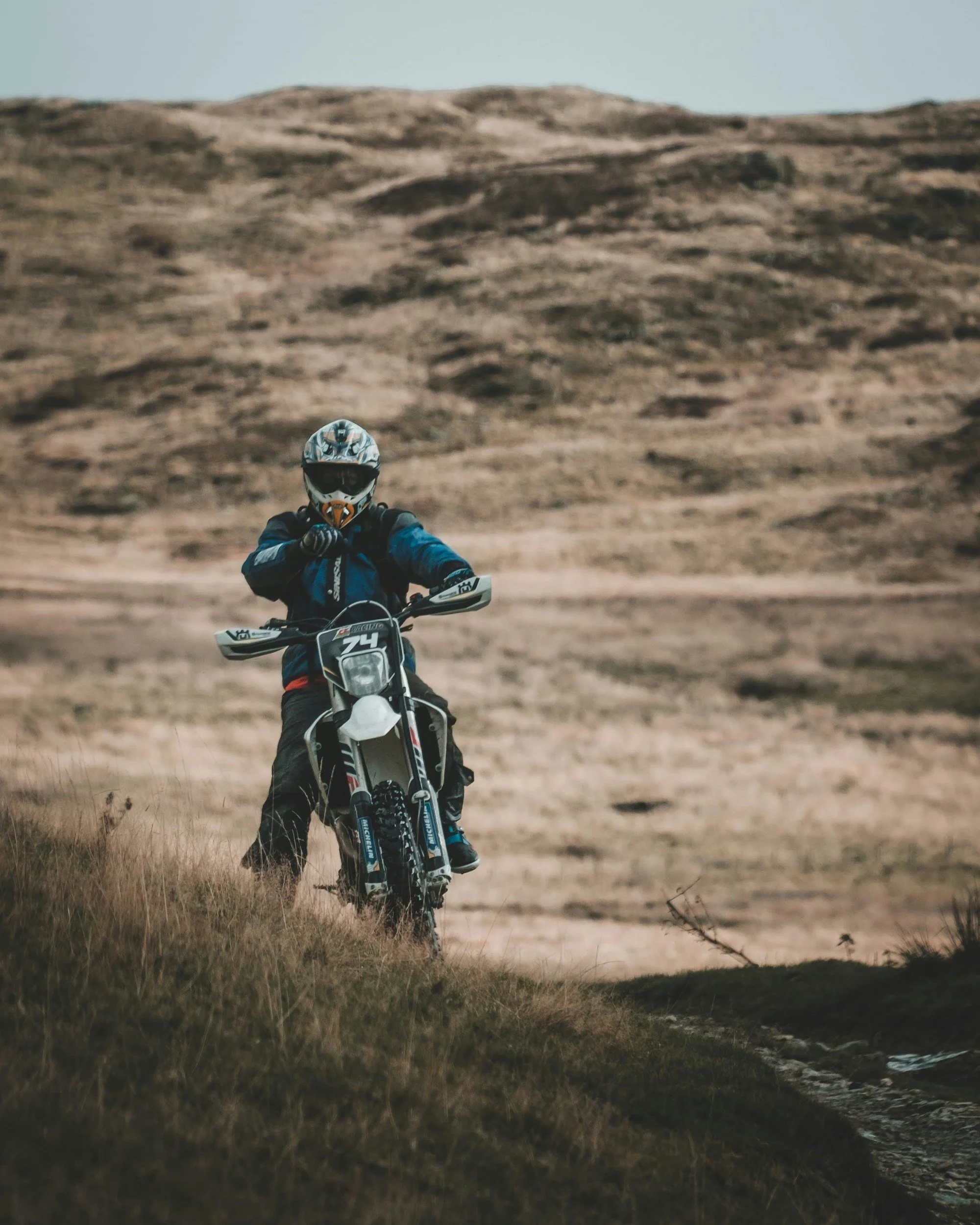 A person riding a dirt bike in an open landscape with dry grass and hills in the background.