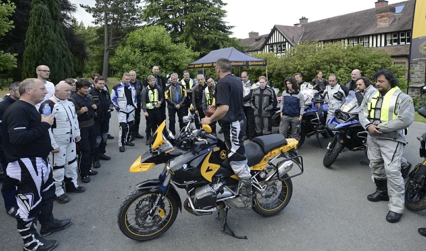 Group of motorcycle riders gathered outdoors around a person sitting on a yellow and black motorcycle, with motorcycles parked nearby on a road lined with trees and buildings in the background.