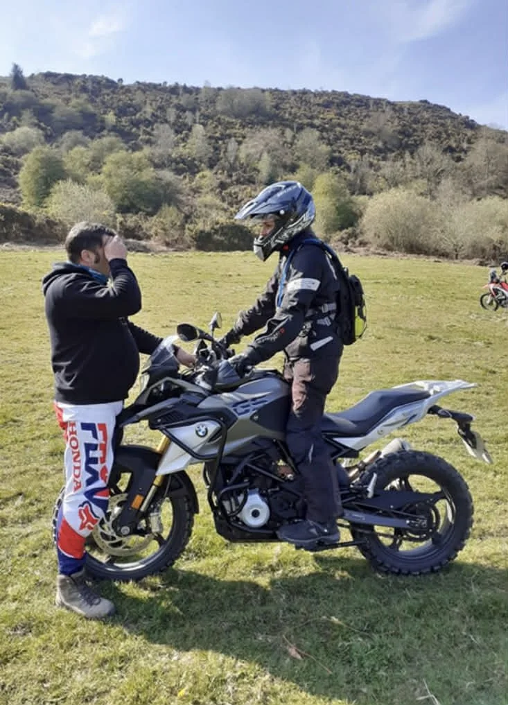 A person in motorcycle gear sitting on a black and gray motorcycle, talking to a young boy in a black hoodie and red and white pants, on a grassy field with hills and trees in the background.