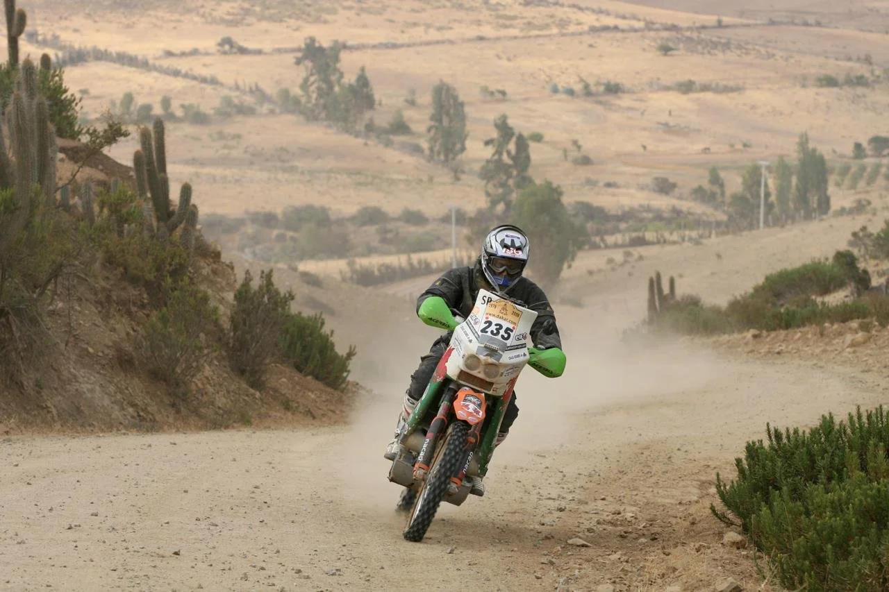 A motorcyclist wearing a helmet and black gear riding a dirt bike on a dusty trail in a desert landscape with cacti and sparse vegetation.