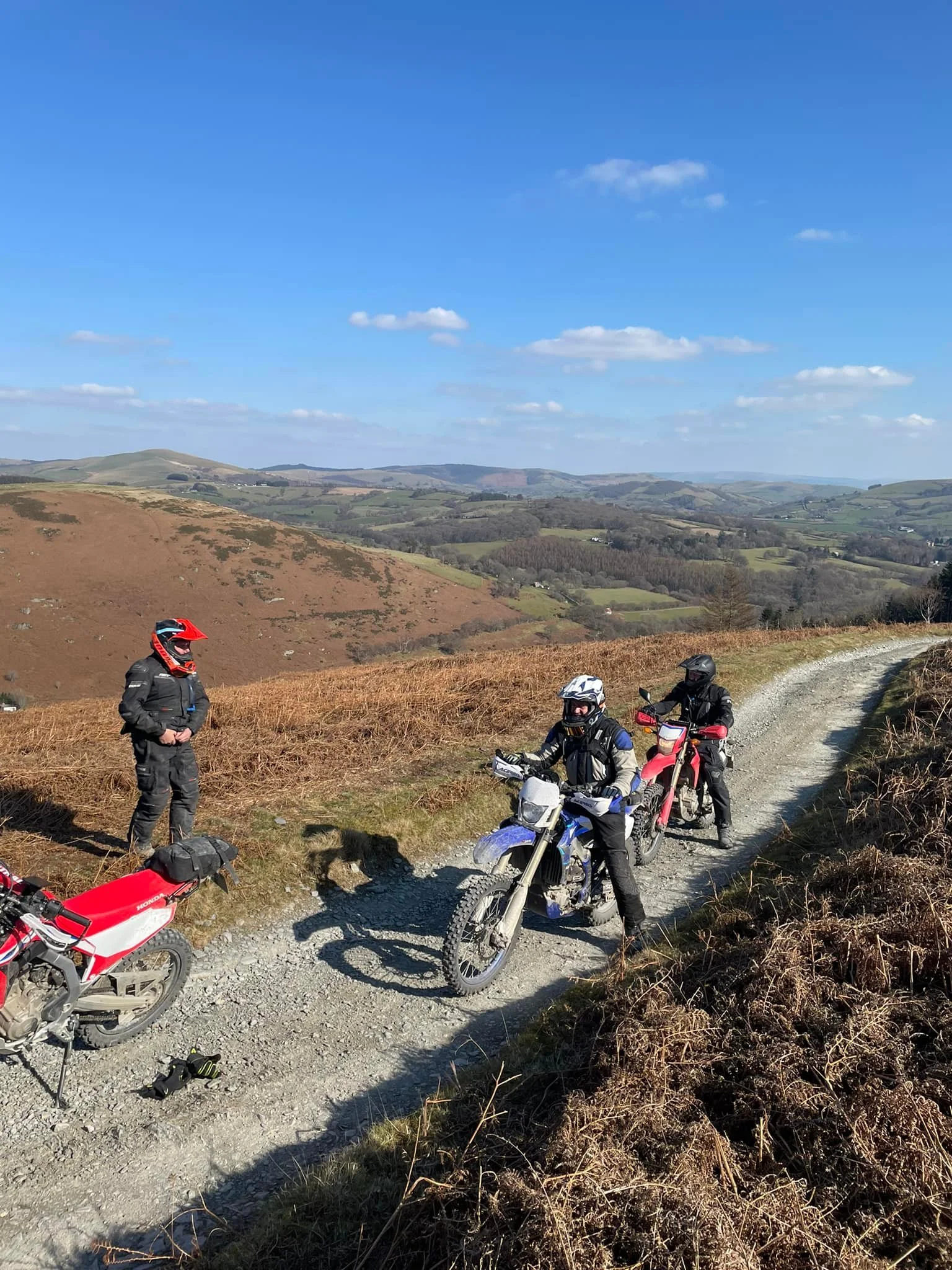 Group of motorcyclists taking a break on a dirt trail in a rural hilly landscape with blue sky and scattered clouds.