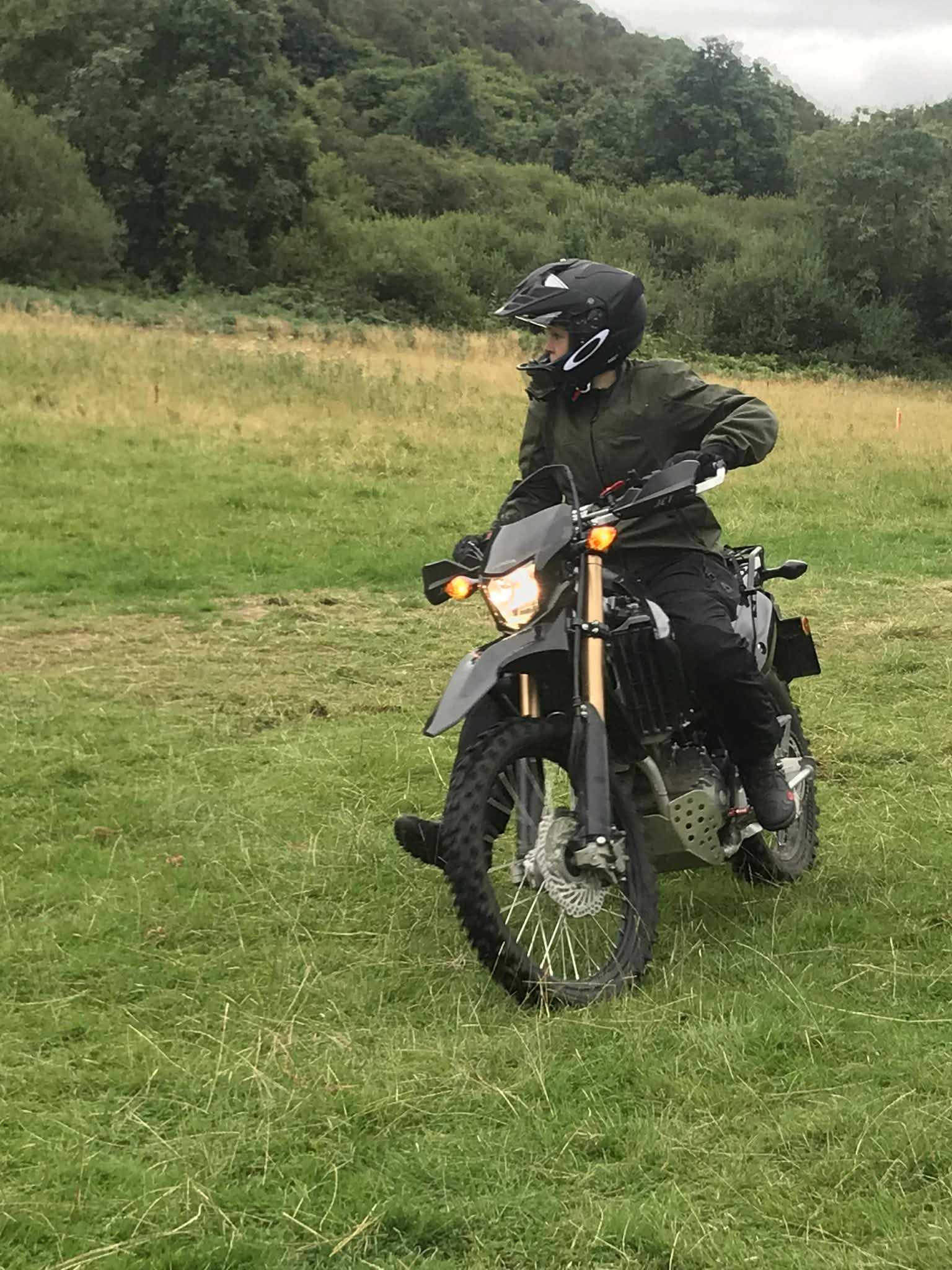 A person wearing a black helmet and dark jacket sitting on a black motorcycle on a grassy field with green trees and hills in the background.