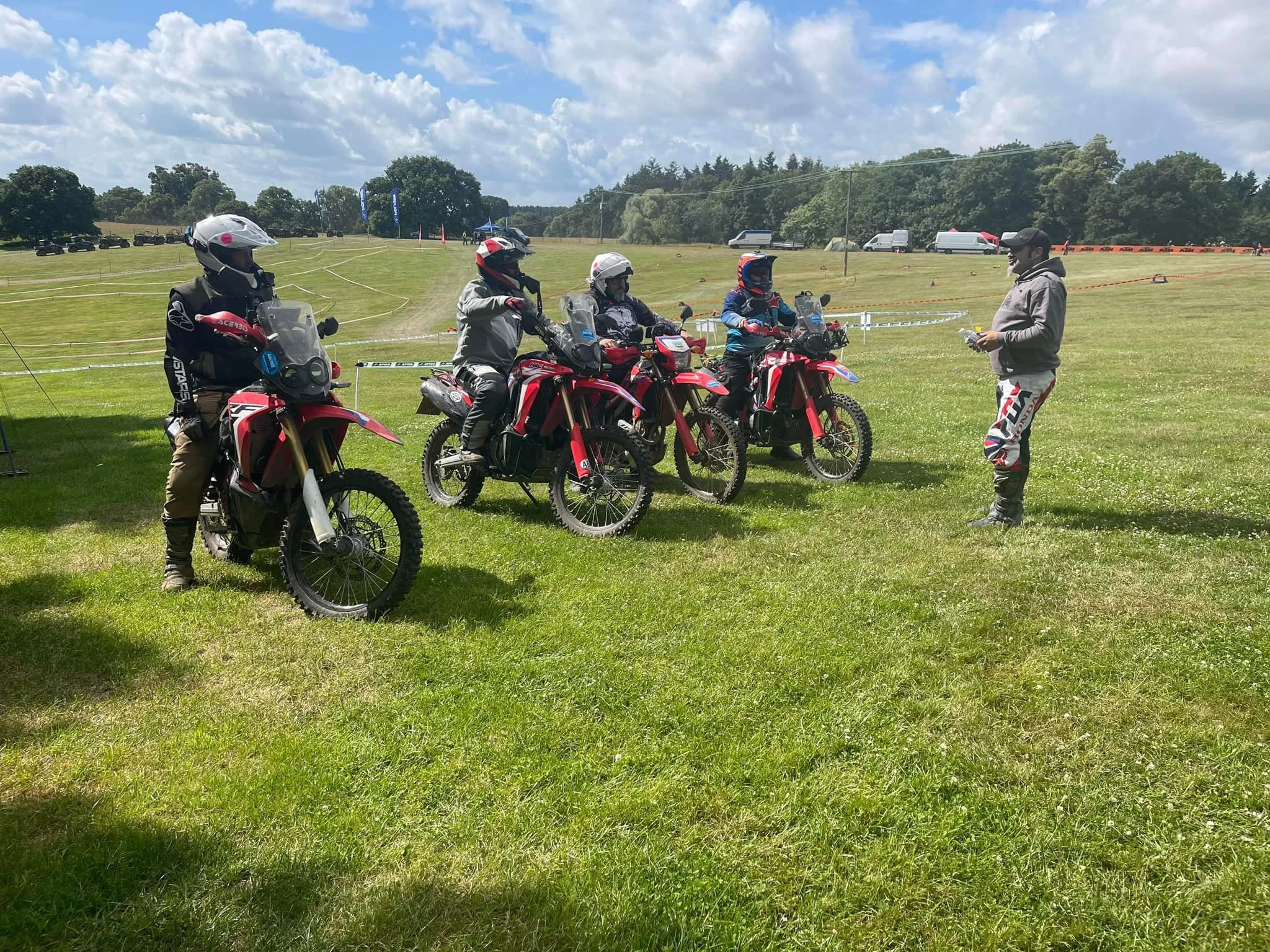 Group of five people with helmets and protective gear sitting on red dirt bikes, listening to a person in a gray hoodie and black cap speaking to them on a green field under a partly cloudy sky.
