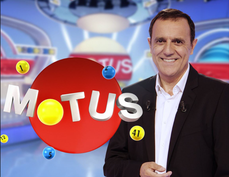 Image d'un homme souriant en costume noir devant un décor de jeu télévisé avec le logo 'Motus' en gros caractères blancs sur un fond rouge et plusieurs balles numérotées
