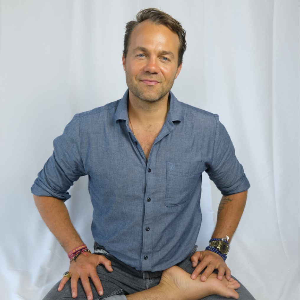 Portrait of a Hatha Yoga and Vajrayana Buddhist teacher seated against a white background, representing embodied practice, clarity, and grounded presence.